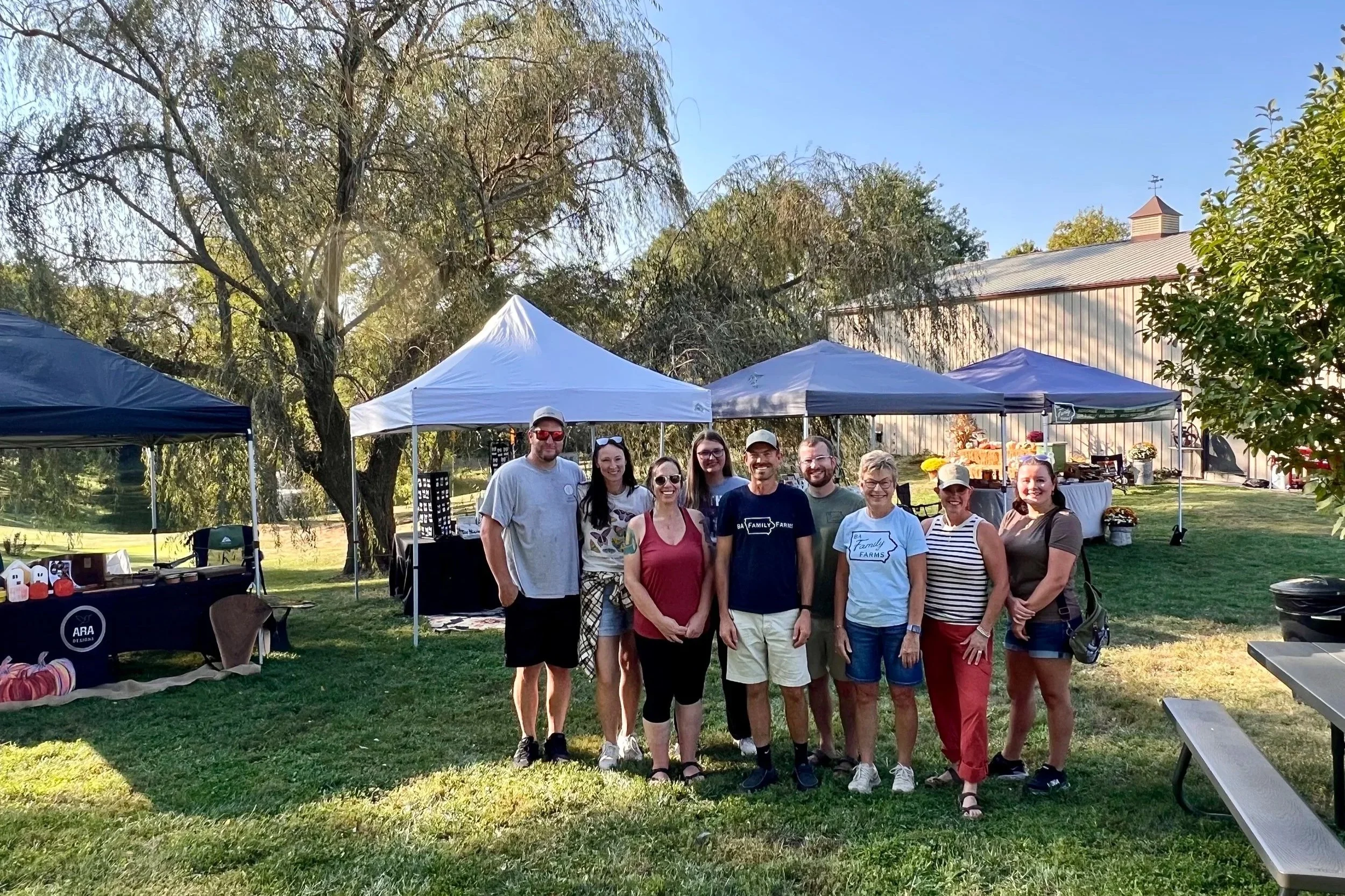 Group of nine people standing together outdoors at a gathering with tents and a barn in the background.