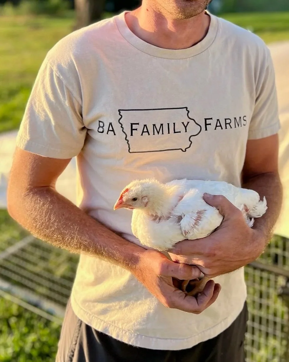 A person holding a young chicken outdoors, wearing a beige T-shirt with 'BA Family Farms' printed on it, near a chicken coop.