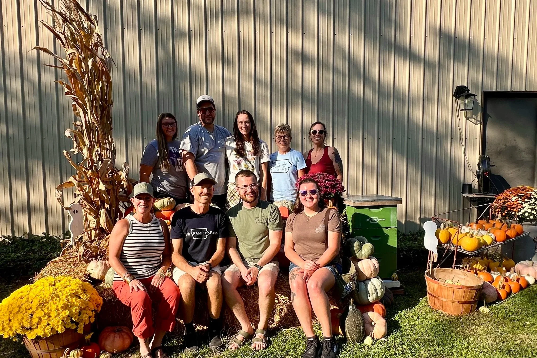 A group people posing outdoors at BA Family Farms during the Backroads & Byways tour, surrounded by fall decorations such as pumpkins, gourds, and mums, with a hay bale and a corn stalk decoration visible.