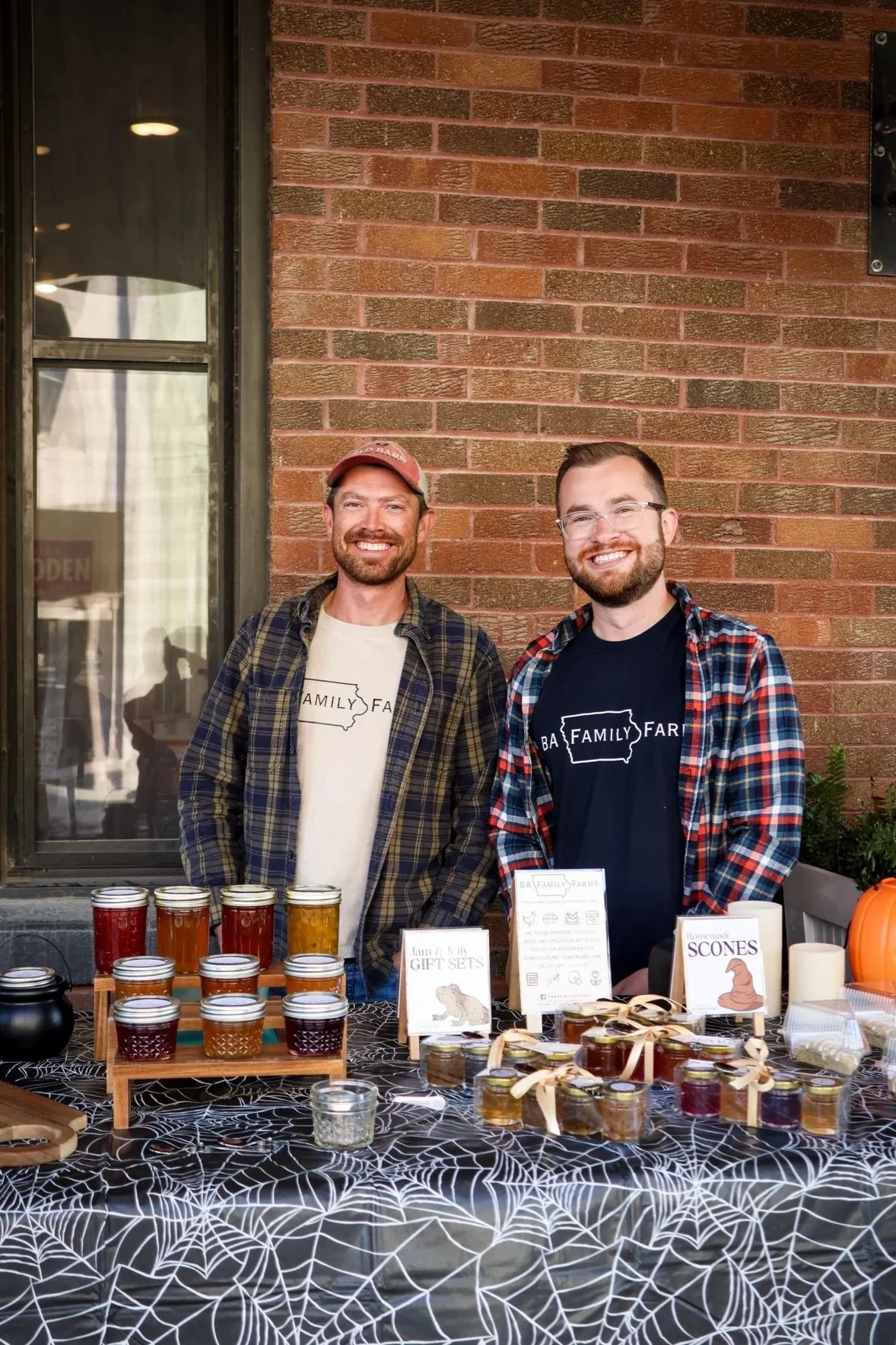 The owners of BA Family Farms stand behind a table with jars of homemade jams and honey, at an event with a brick wall and window in the background.