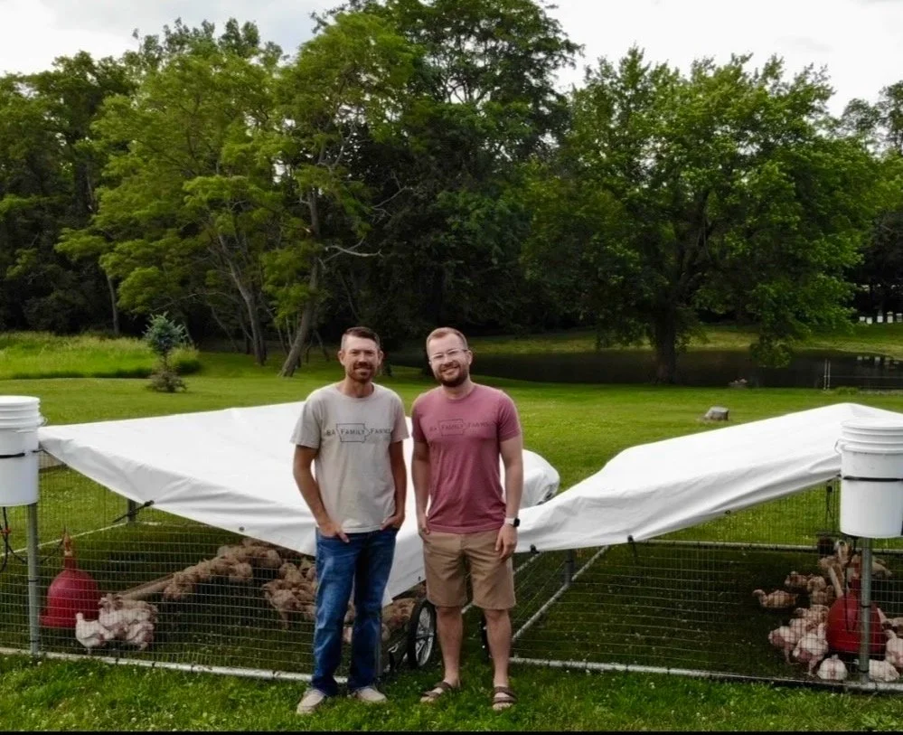The owners of BA Family Farms stand in front of two chicken tractors, in a grassy area with trees and a pond in the background.