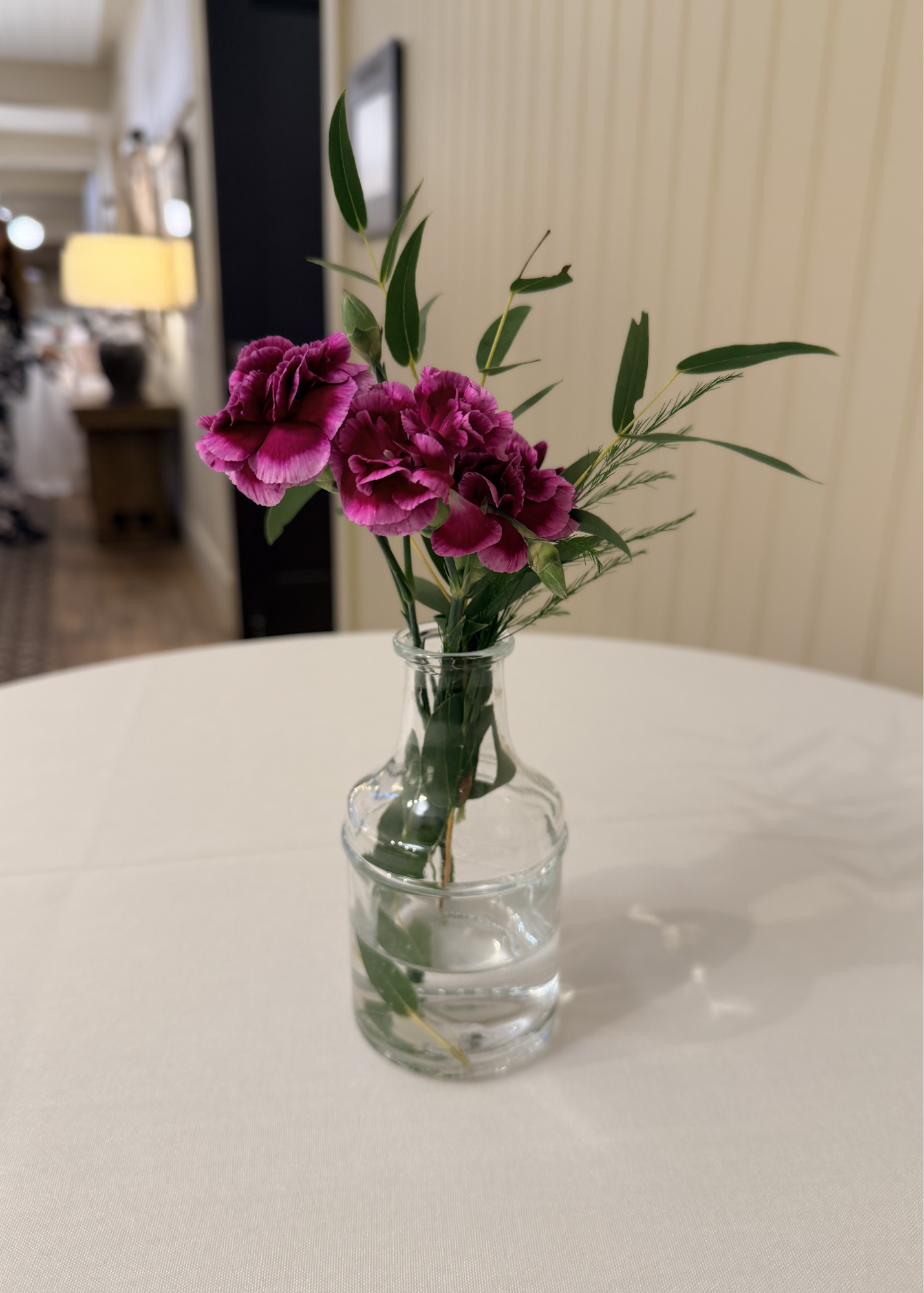 Pink and purple carnations in a clear glass vase on a white table in a room with beige walls and background furniture.