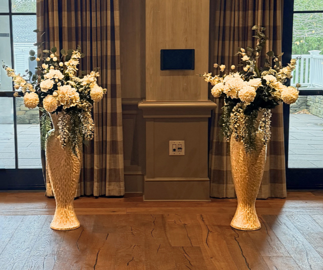 Two large gold vases filled with white flowers and greenery are on either side of a wall with curtains, with a small wall-mounted speaker and electrical outlet between them, in front of glass doors leading outside.