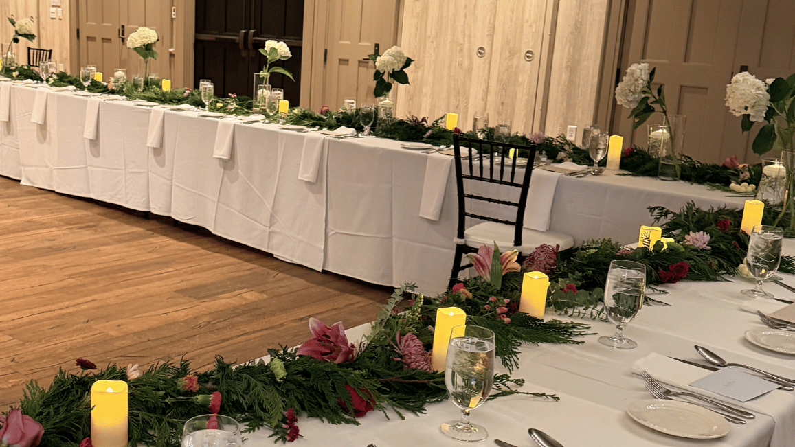 Long banquet table decorated with white floral arrangements, candles, greenery, and pink flowers, set for a formal event in a banquet hall.