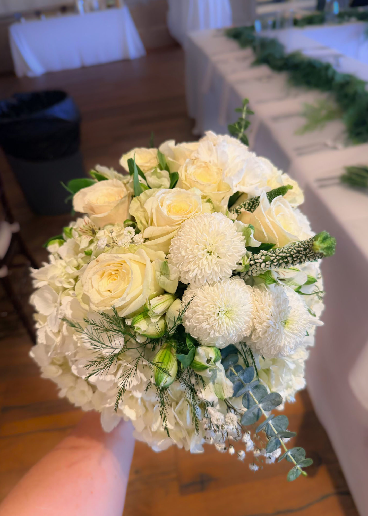 A bouquet of white roses, chrysanthemums, and greenery held by a person in a room with decorated tables in the background.