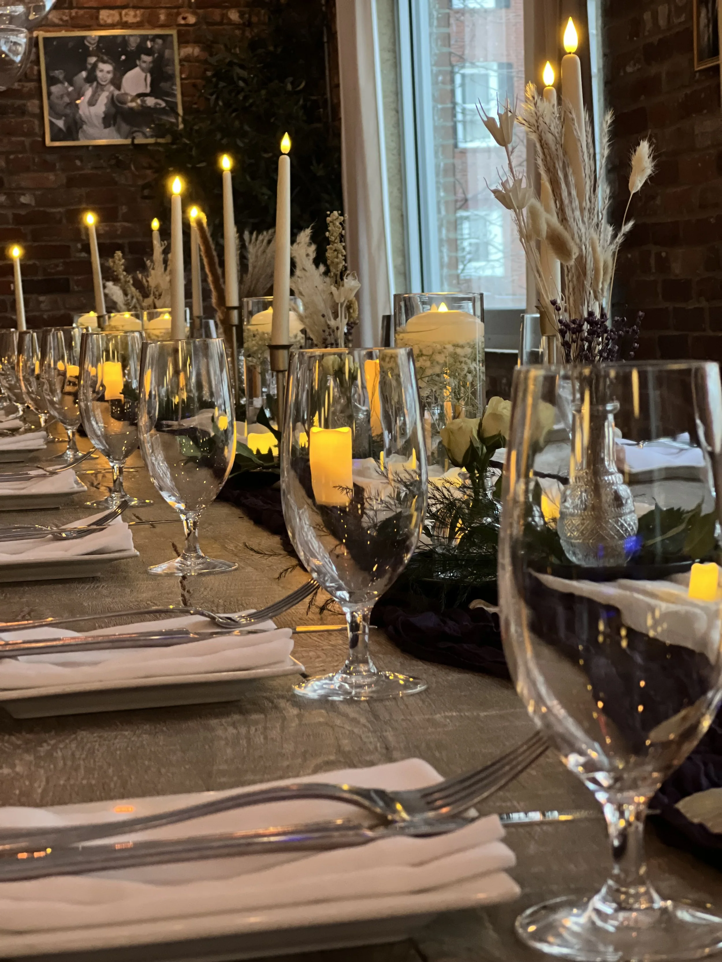 Elegant dining table set with wine glasses, white napkins, and silverware, decorated with candles and floral arrangements, in a cozy, warmly lit room with brick walls and a window.