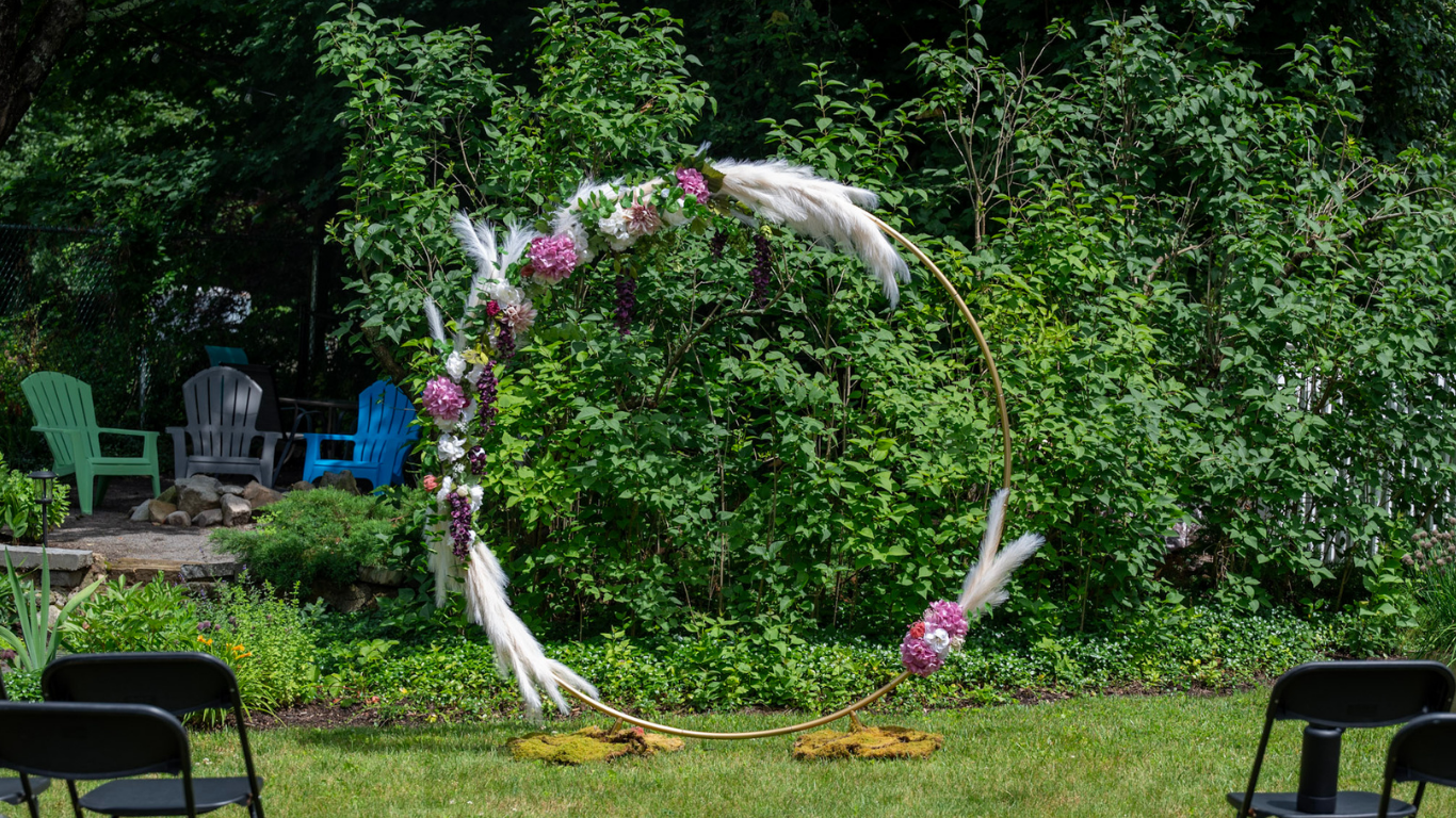 Outdoor garden scene with a circular wedding arch decorated with pink and white flowers, white feathers, and greenery. There are three chairs, one black, one green, and one blue, arranged around the arch with a lush green background of trees and bush