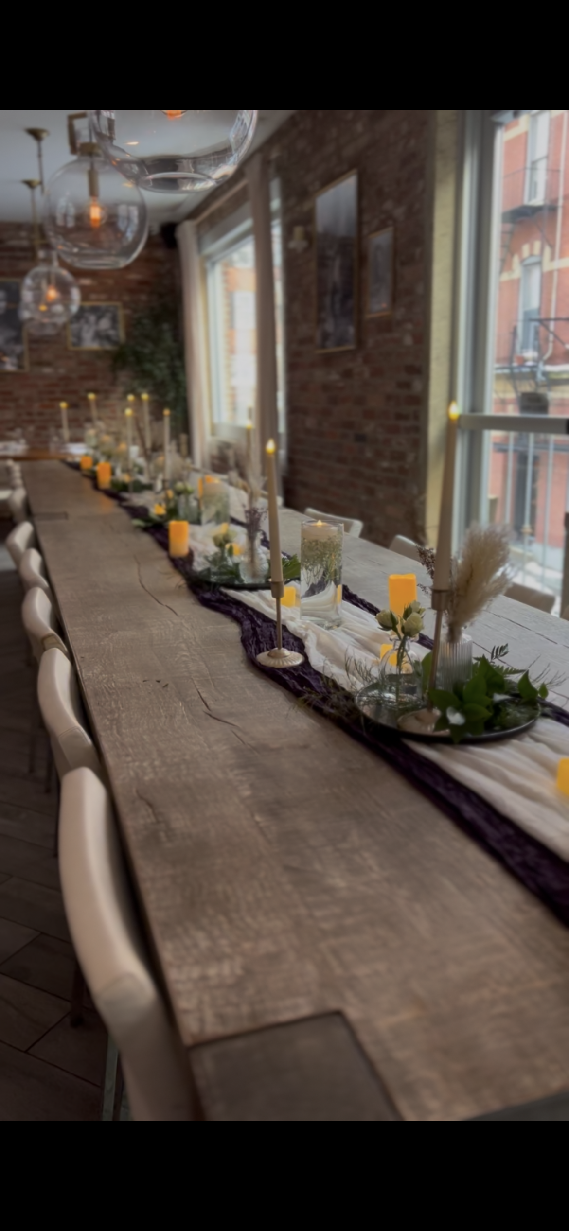 A decorated banquet table set for a gathering with candles, vases, and greenery in a room with exposed brick walls and large windows.