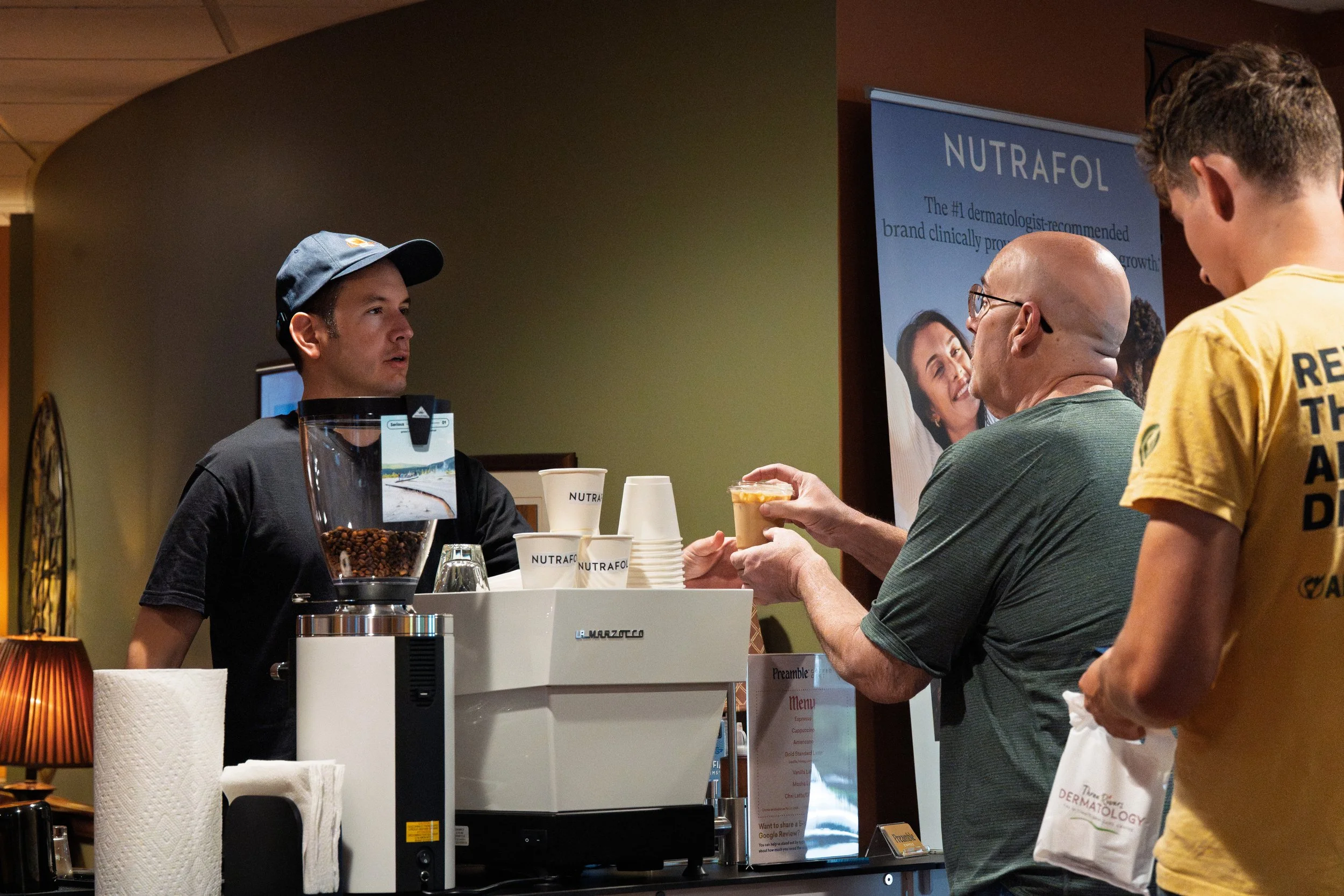 Barista Mark Pape of Preamble Coffee Cart Catering in Fort Wayne, Indiana happily serving an iced latte made with premium espresso and syrups to an excited customer at a dermatology event with other customers ready to order.