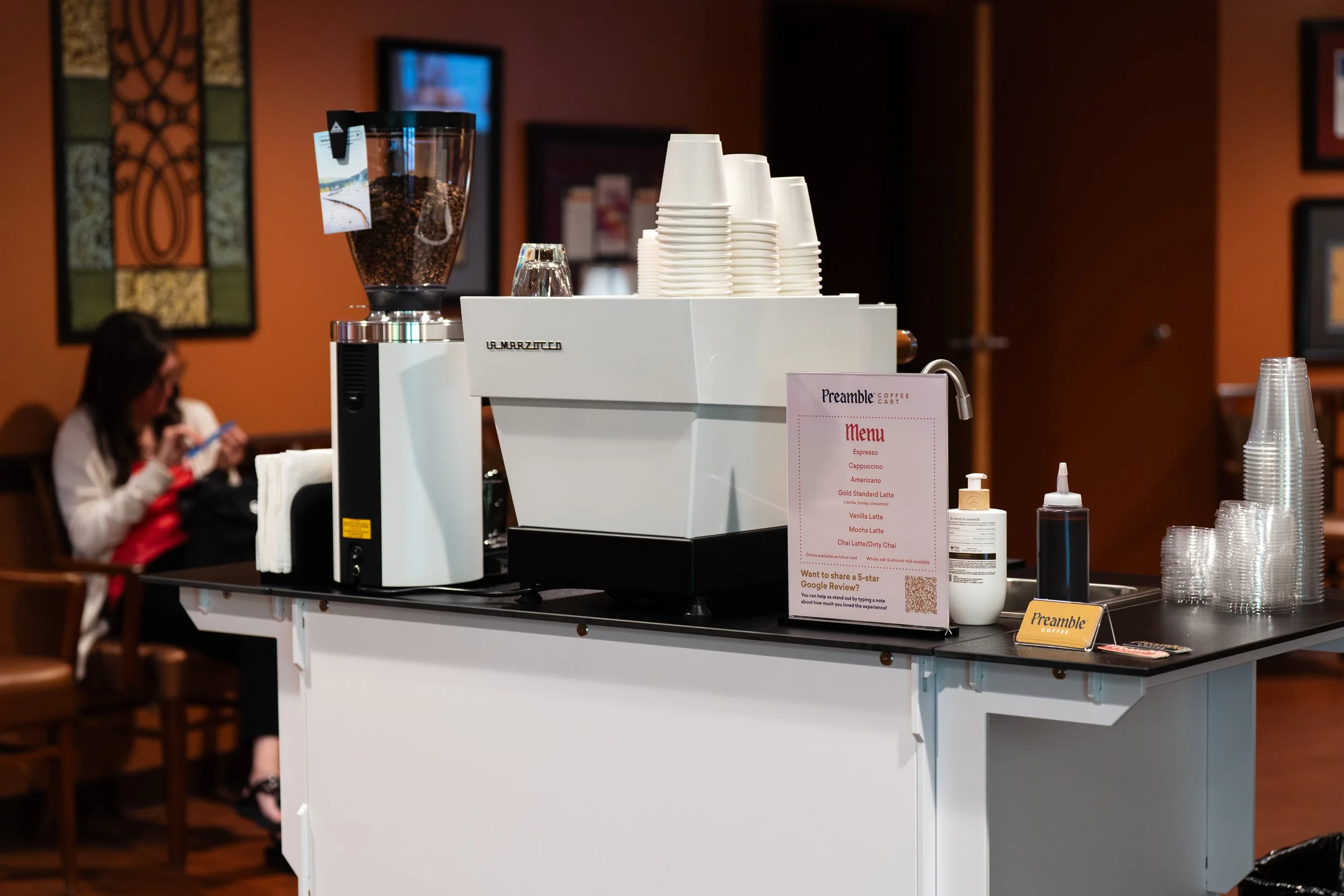 Preamble Coffee Cart Catering in Fort Wayne, Indiana ready to serve premium espresso drinks using the best equipment to patients at a dermatology office with a customer waiting for her appointment nearby.