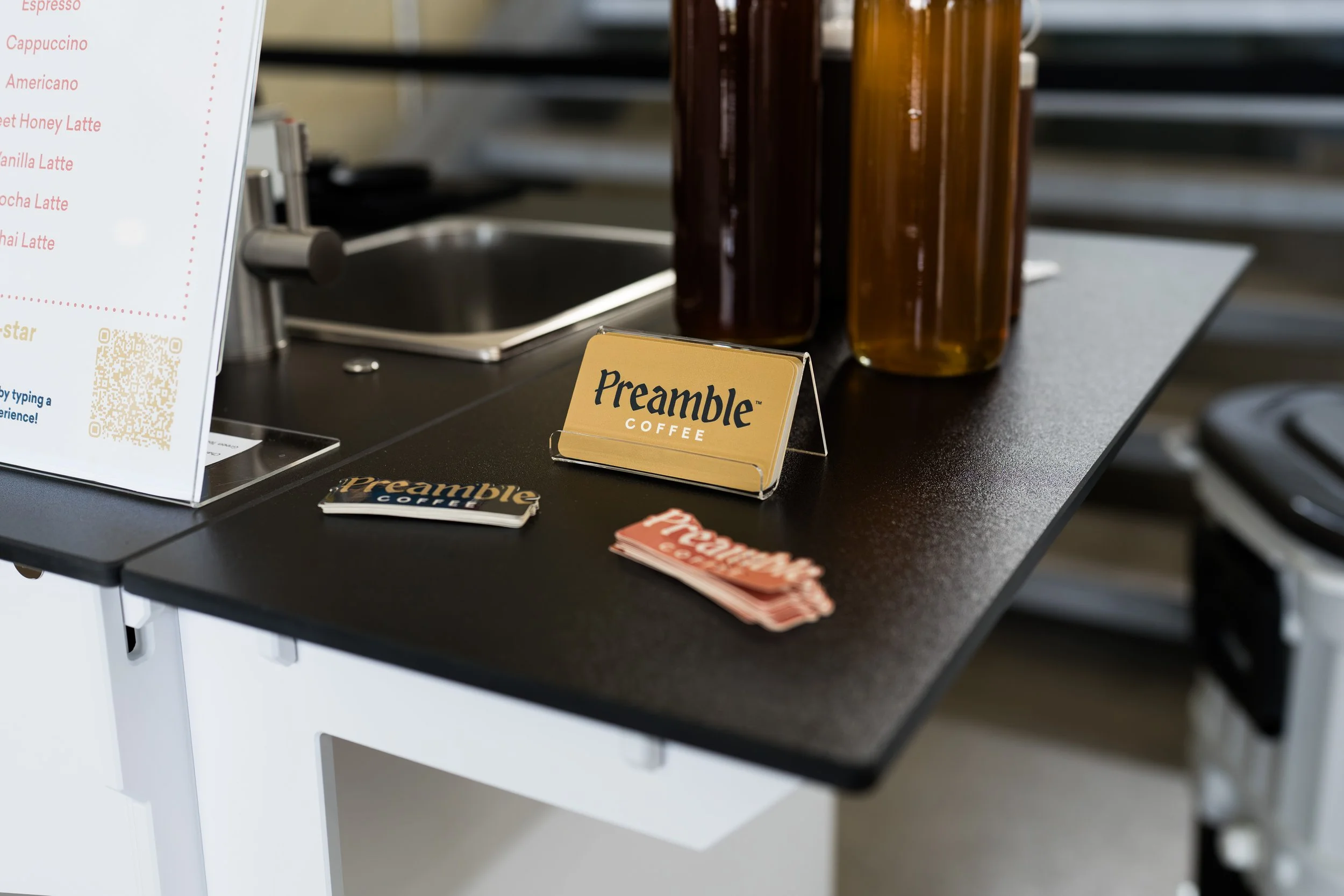 A black countertop with a yellow sign that reads 'Preamble Coffee,' acrylic stand for the sign, a small stack of Preamble Coffee cards, and several brown and orange bottles or containers are on the counter. To the left, part of a white menu board with red text is visible.