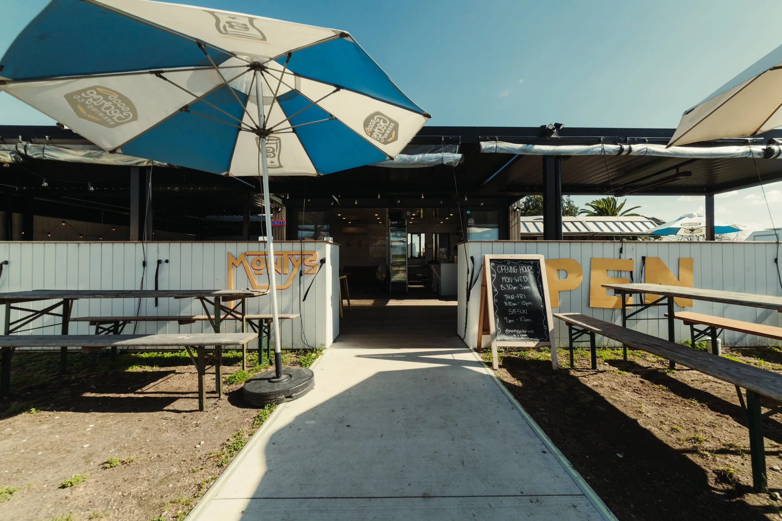 Outdoor seating area of a restaurant with picnic tables and umbrellas in front of a white fence, open entrance to the restaurant visible, and a chalkboard sign with opening hours.