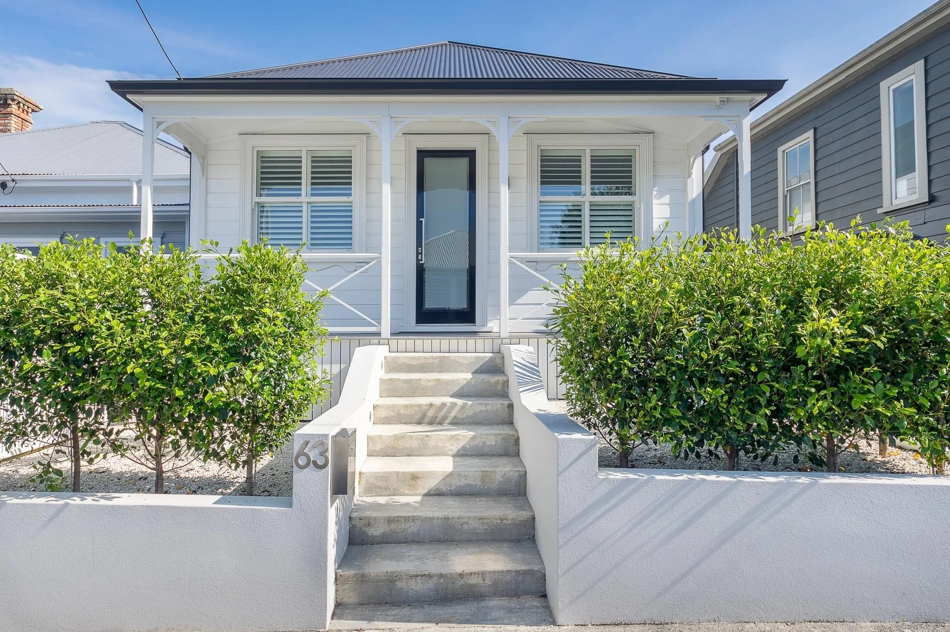 Front view of a white house with a porch, stairs, and green bushes, on a sunny day.