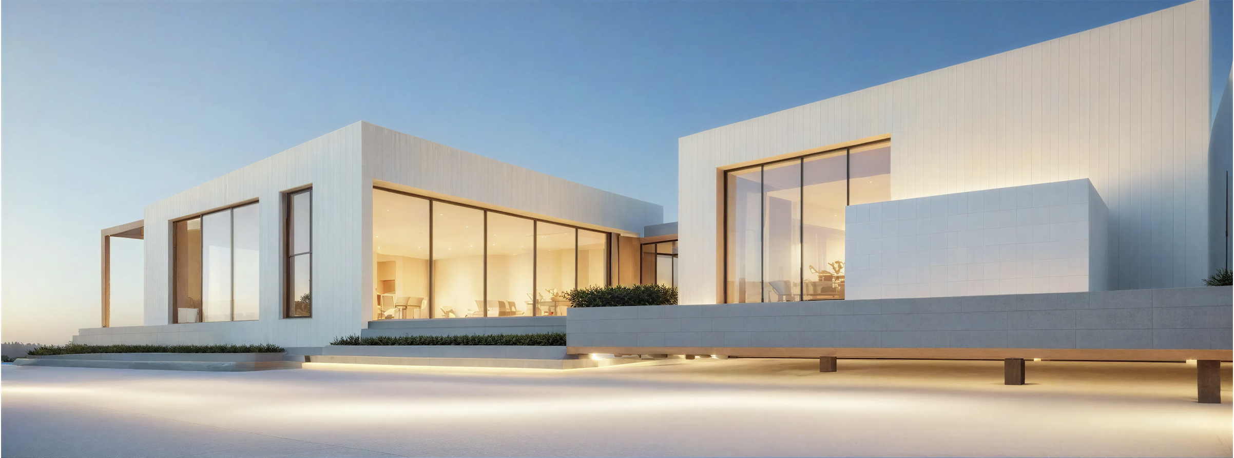 Modern white building with large glass windows at dusk, illuminated from inside, with a smooth concrete foreground and landscaped greenery.