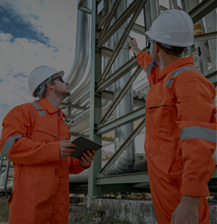 Two workers in orange safety uniforms and white helmets inspecting industrial equipment. One holds a tablet and the other points with a tool.