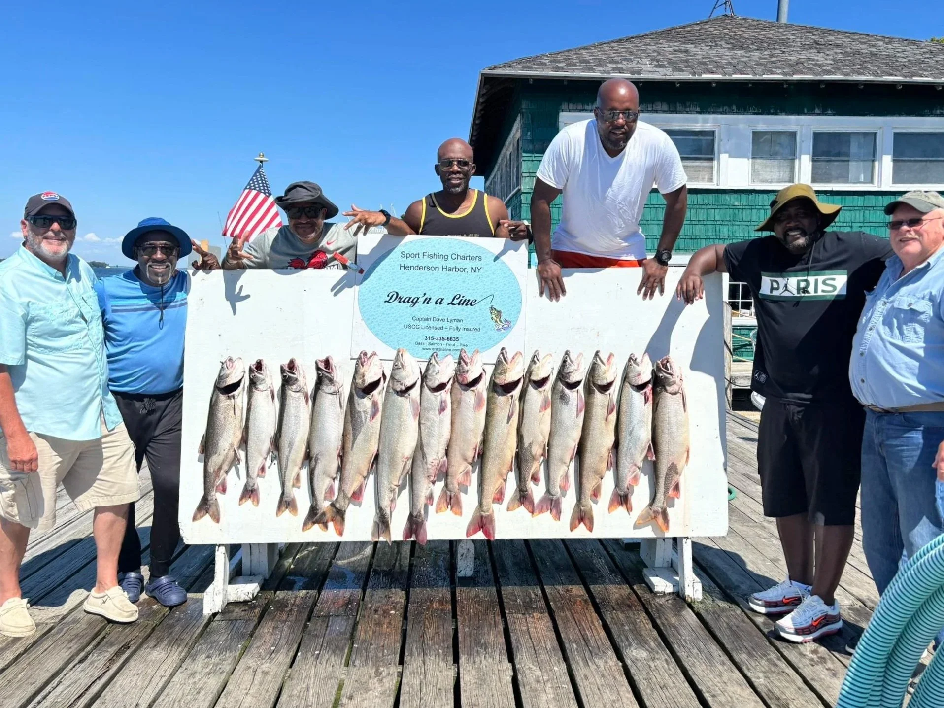 Group of men standing on a wooden dock with a large display board of caught fish, posing with the catch, under blue sky and building in background.