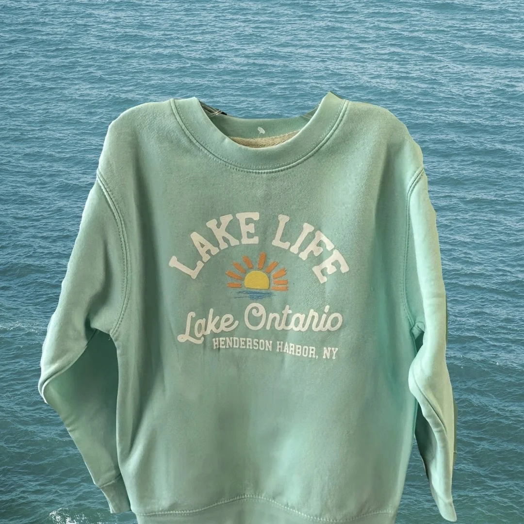 A light green sweatshirt with the text 'Lake Life' and an illustration of a setting sun over water, with additional text 'Lake Ontario Henderson Harbor, NY', against a background of water.