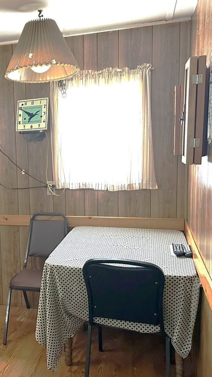 A small wood-paneled dining area with a table covered in a polka dot tablecloth, two chairs, a window with sheer curtains, a hanging lamp, a clock on the wall, and a remote control on the table.