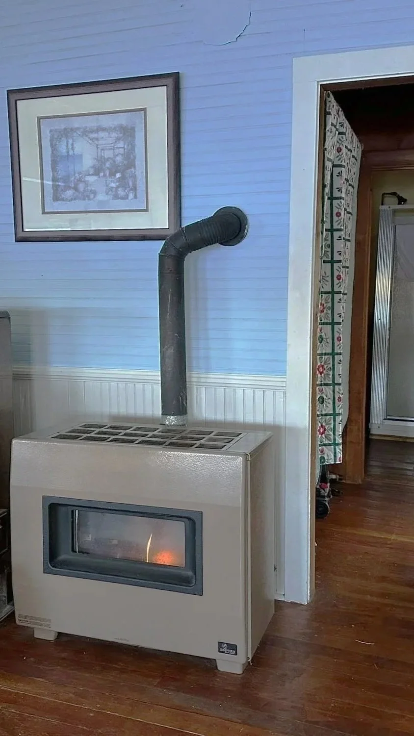 A vintage wood stove in a room with wood flooring and light blue paneling walls. There is a framed picture above it and a black pipe extending from the stove to the wall. Part of a doorway with patterned curtains is visible to the right.