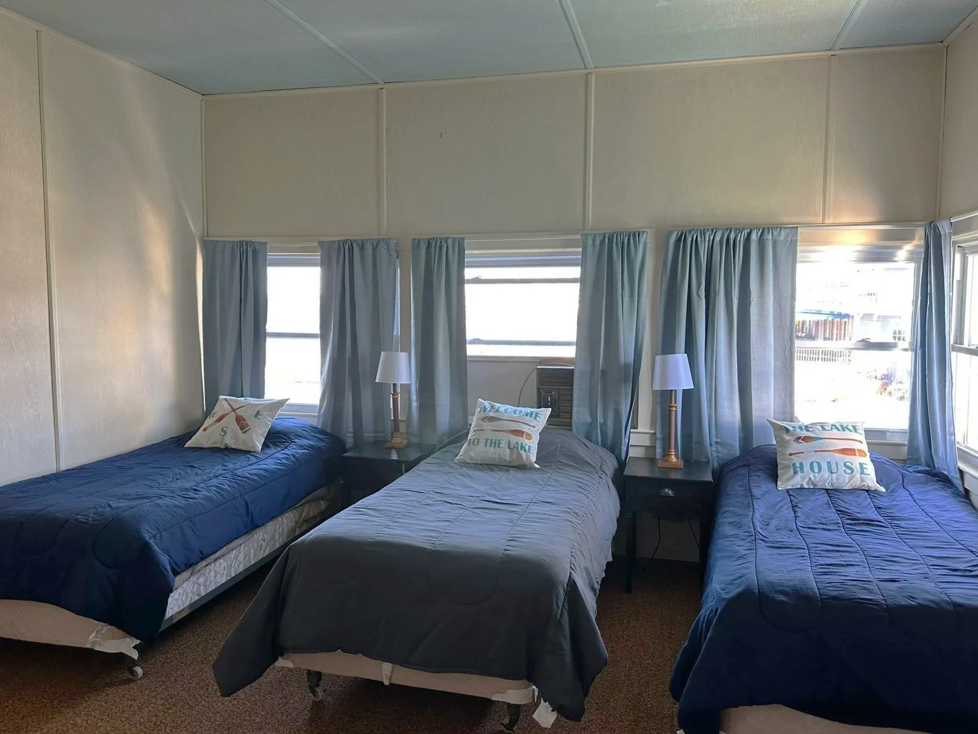 Three beds with blue and gray bedding in a room with windows, blue curtains, and nightstands with lamps.