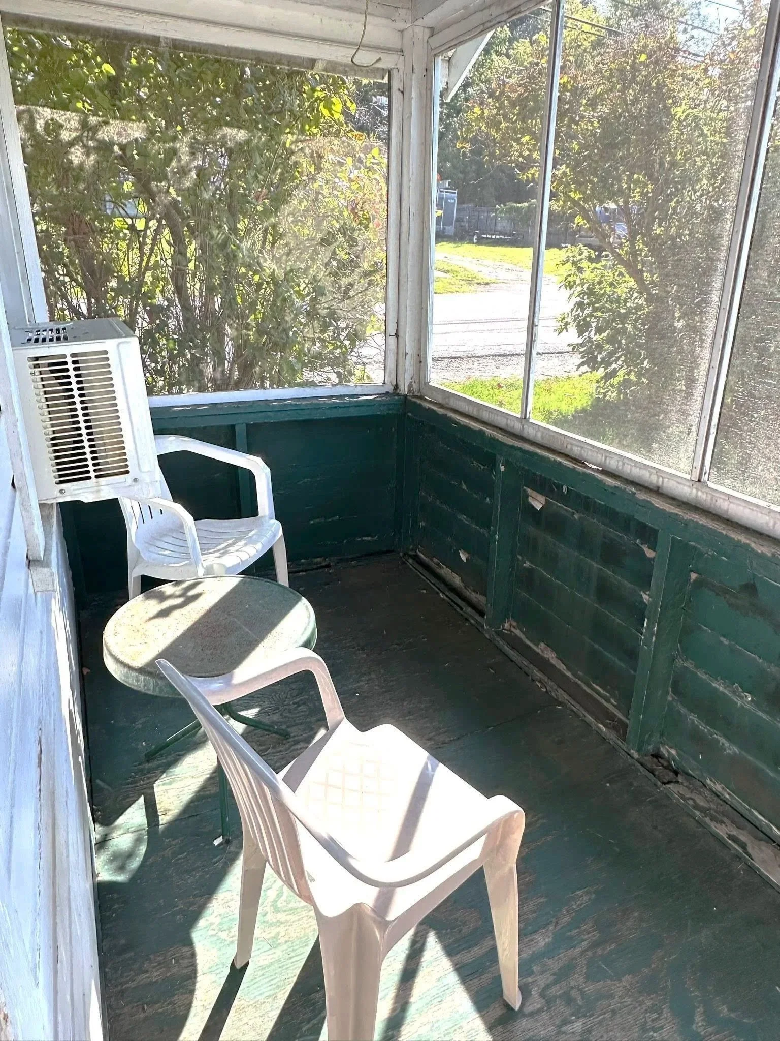 A screened porch with two white plastic chairs and a small round table, weathered green painted walls, a white fan, and surrounding greenery outside.