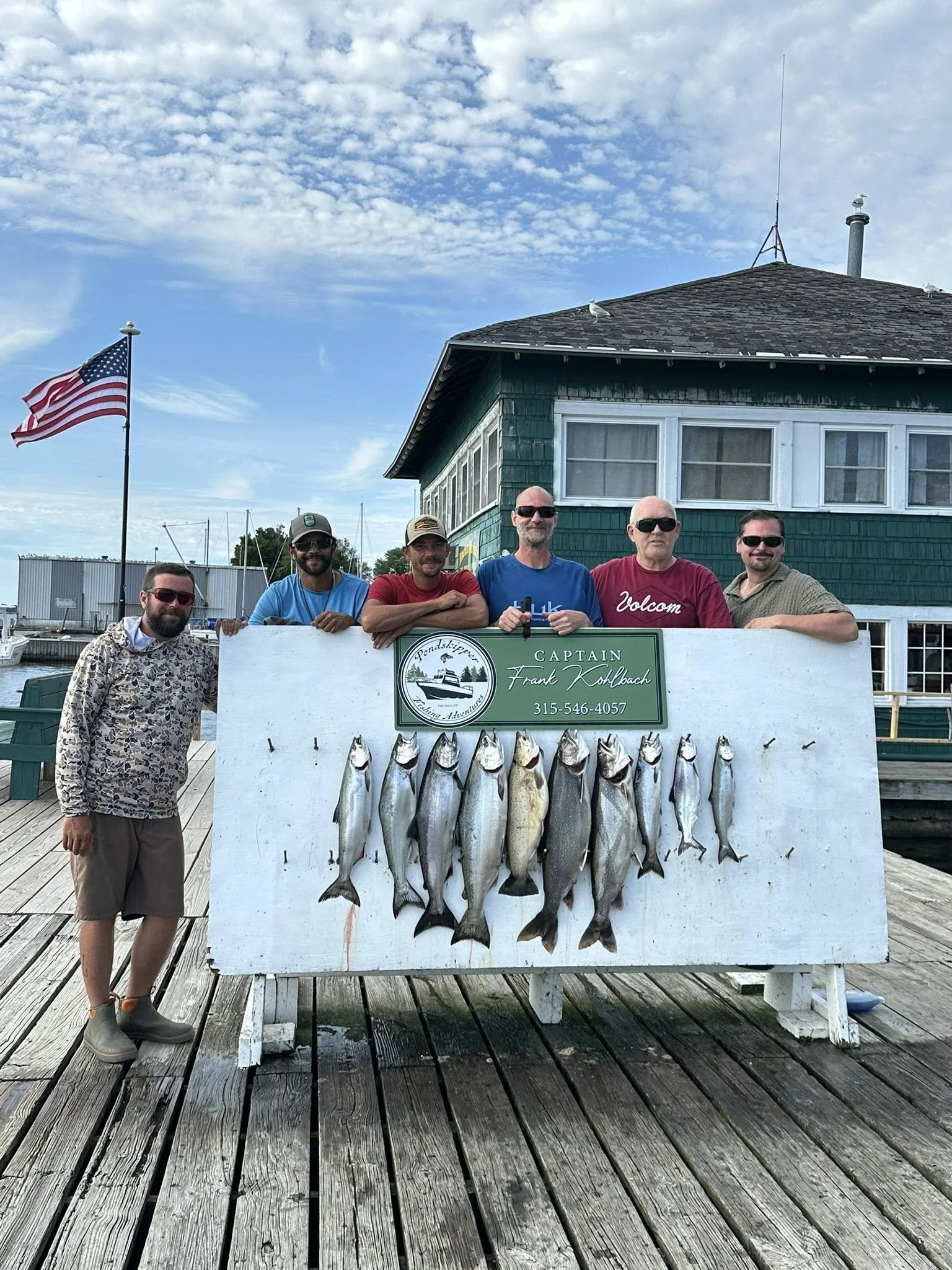 Six men standing and leaning on a white board with fish hung on it, on a wooden dock near a green building; there's an American flag in the background, and a blue sky with scattered clouds.
