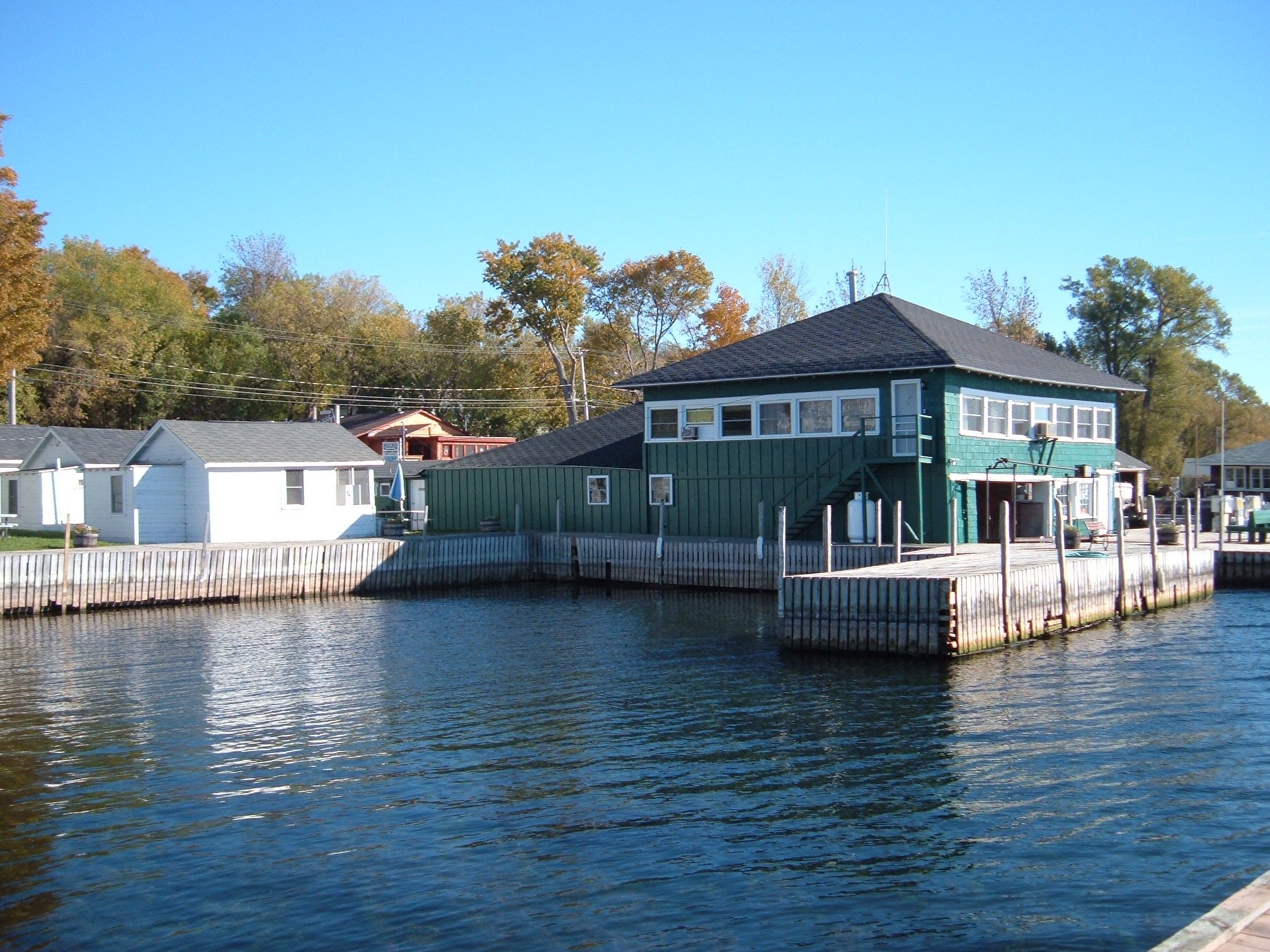 A teal and gray boathouse on a waterway, with neighboring white boathouses, trees, and a clear blue sky in the background.