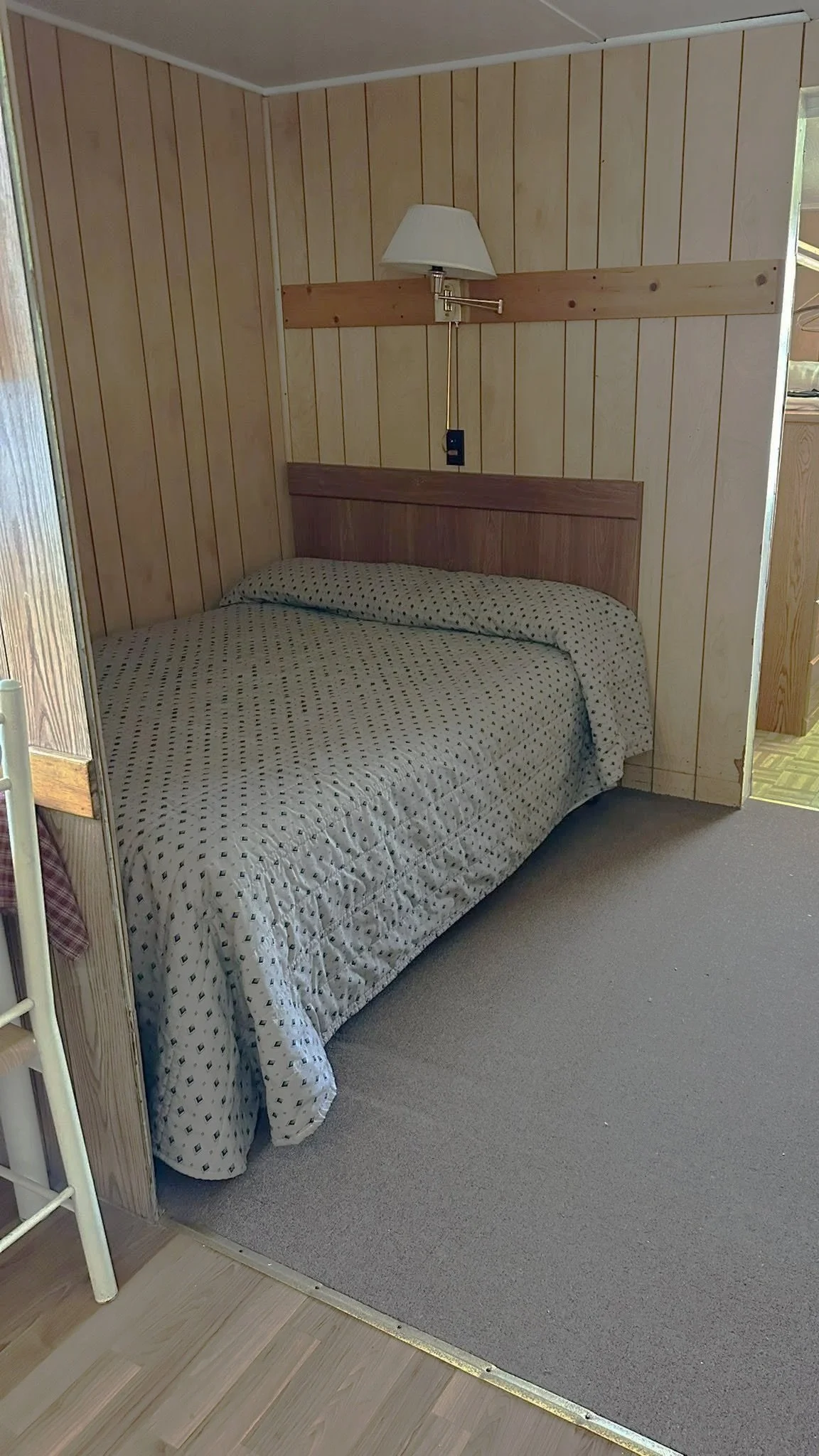 A bedroom with a bed covered in a patterned bedspread. Wood panel walls and a small window are visible, with a wall-mounted reading lamp above the headboard.