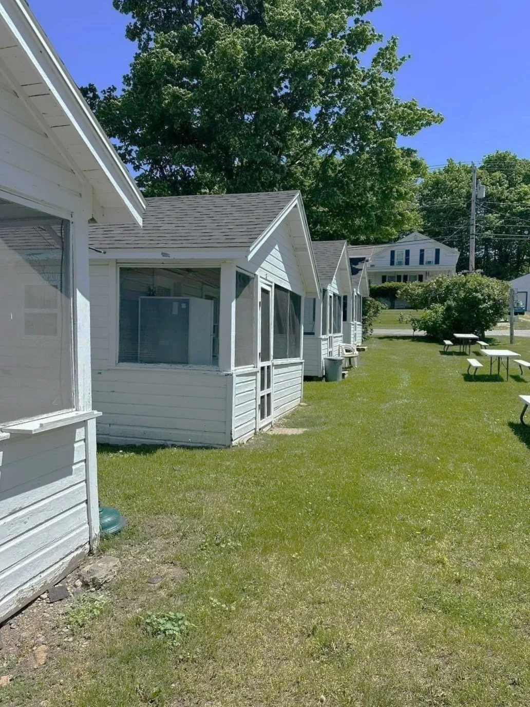 Line of small white cabins with screened porches, green grass, picnic tables, trees, and a clear blue sky.