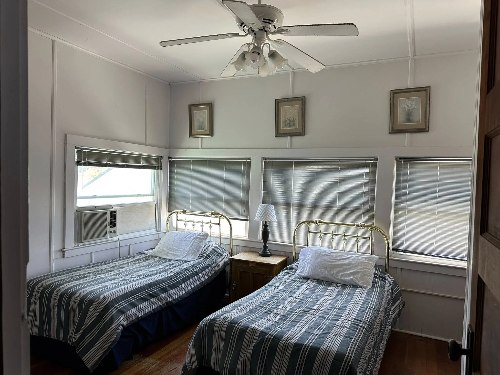 A bedroom with two twin beds with gold metal headboards and striped bedding, a wooden nightstand with a lamp between the beds, three framed pictures of flowers on the wall, a ceiling fan, and windows with blinds and an air conditioning unit.