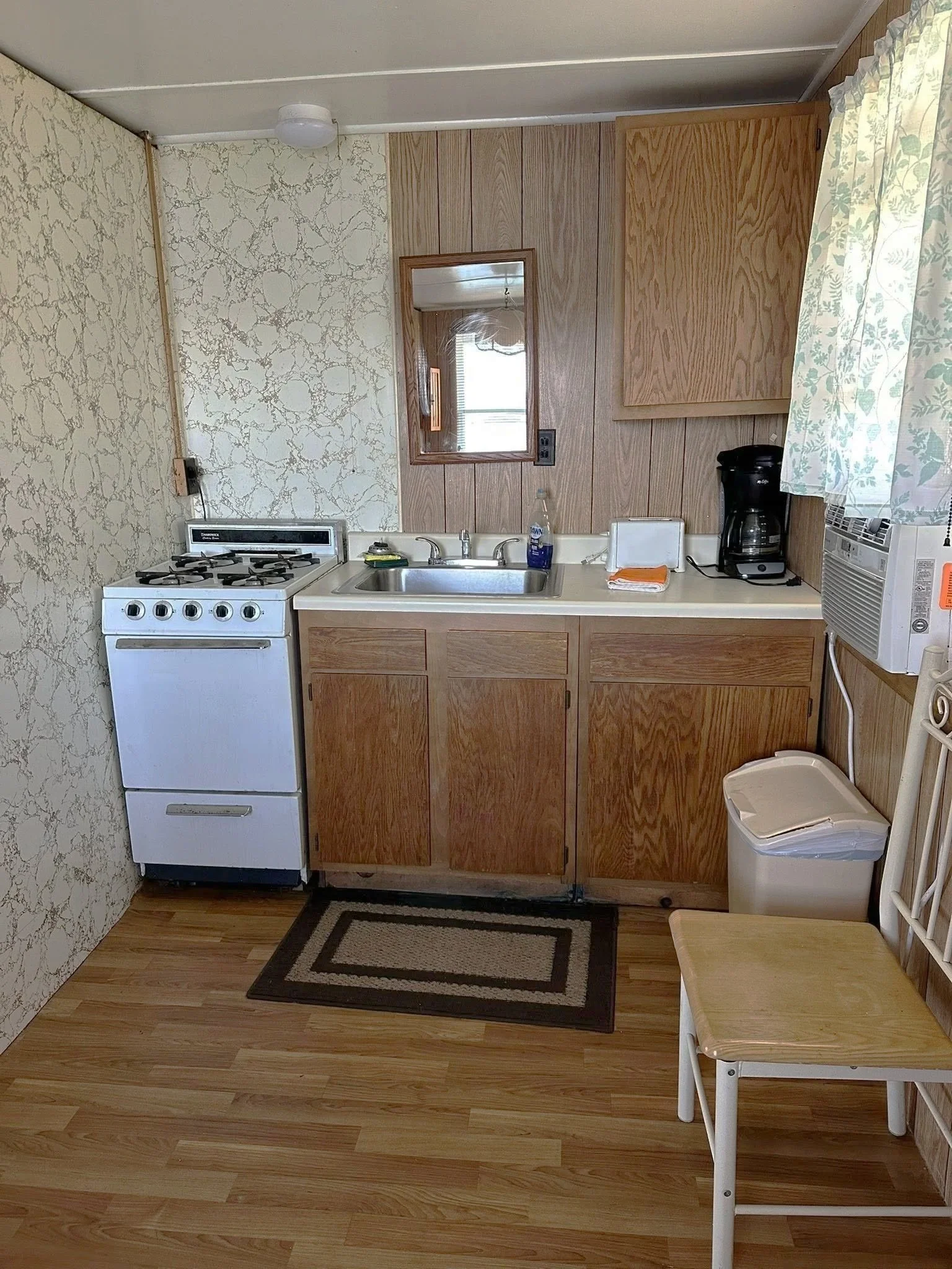 A small kitchenette with a white stove, a stainless steel sink, wooden cabinets, a coffee maker, a window air conditioning unit, a trash can, a wooden chair, and a small rug on a light wood floor.