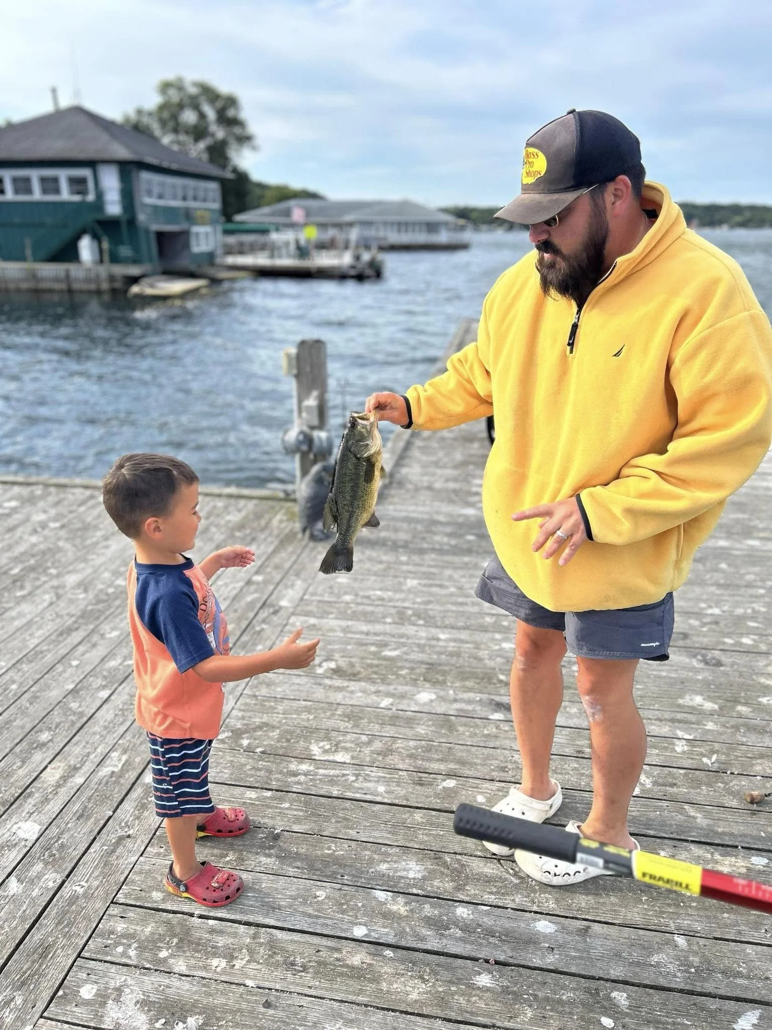 A man in a yellow jacket and cap holding a fish while a young boy in a blue and orange shirt and striped shorts looks on on a wooden dock, with water and houses in the background.
