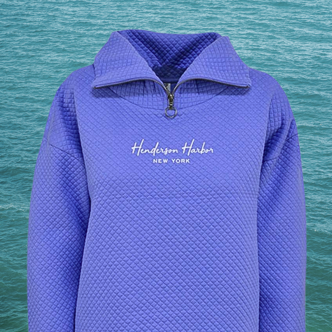 Purple quilted jacket with a quarter-zip collar and white text reading 'Henderson Harbor NEW YORK' on the chest, set against a background of water.