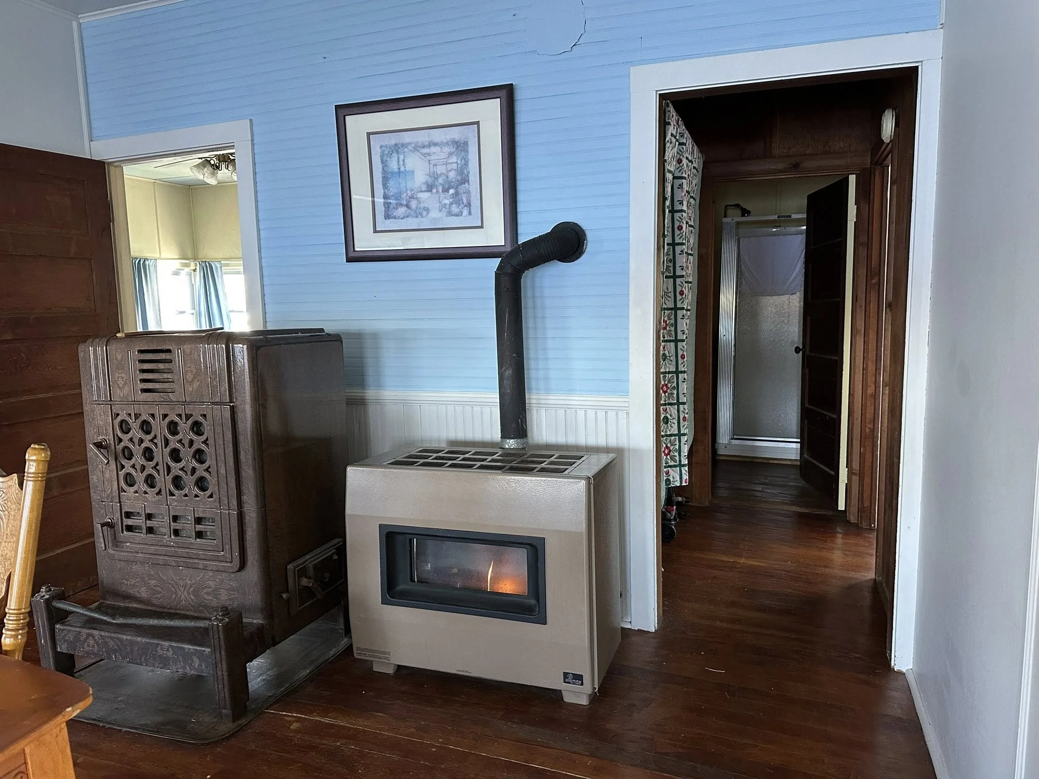 Living room with a wood stove, a vintage heater, a framed picture on a light blue wall, a doorway with a floral curtain leading to a bathroom with a shower, and hardwood floors.