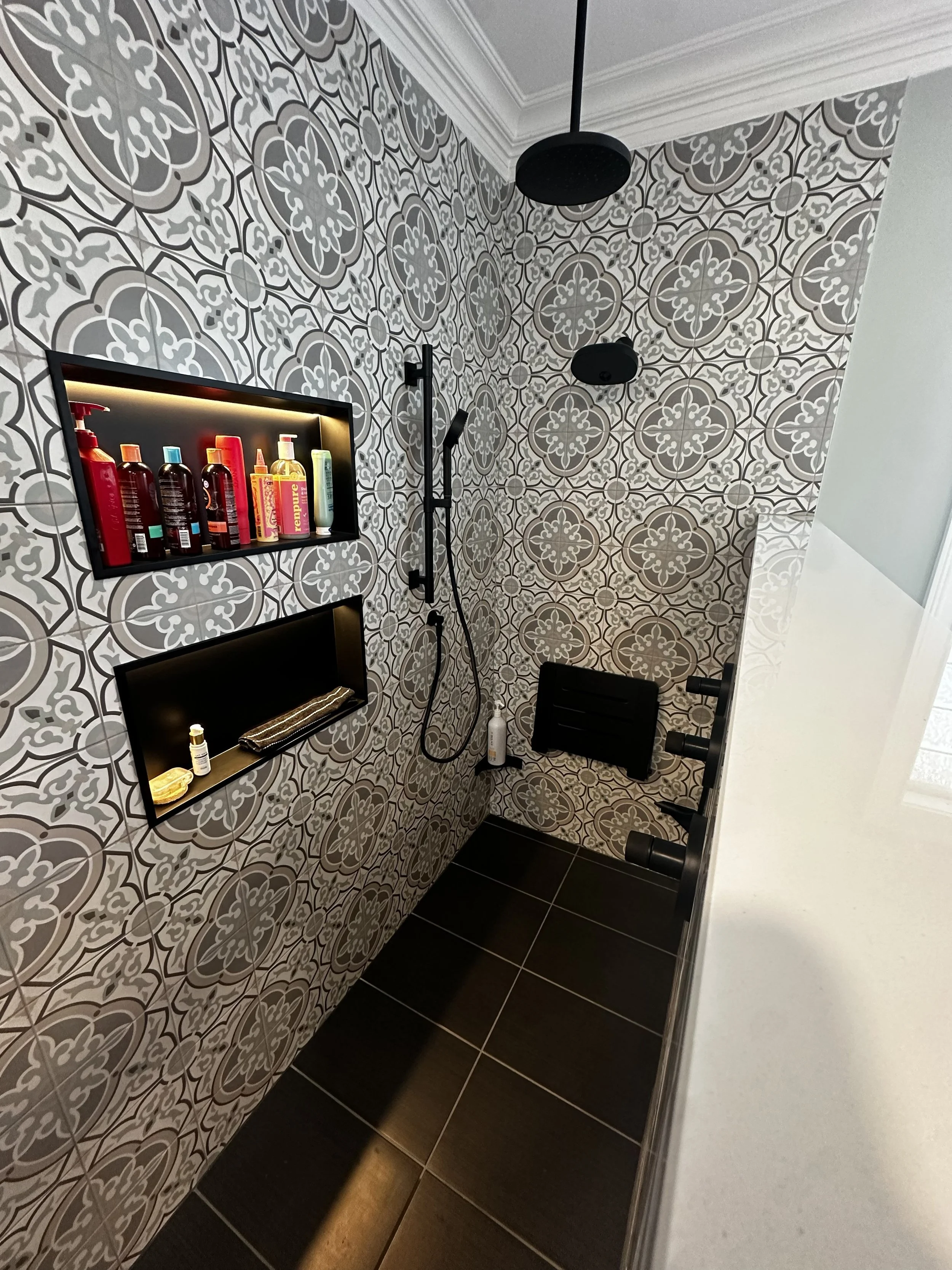 A modern shower with patterned tiles on the walls, black fixtures, shelves with shampoo and toiletries, a handheld showerhead, a wall soap dish, and dark tiled flooring.