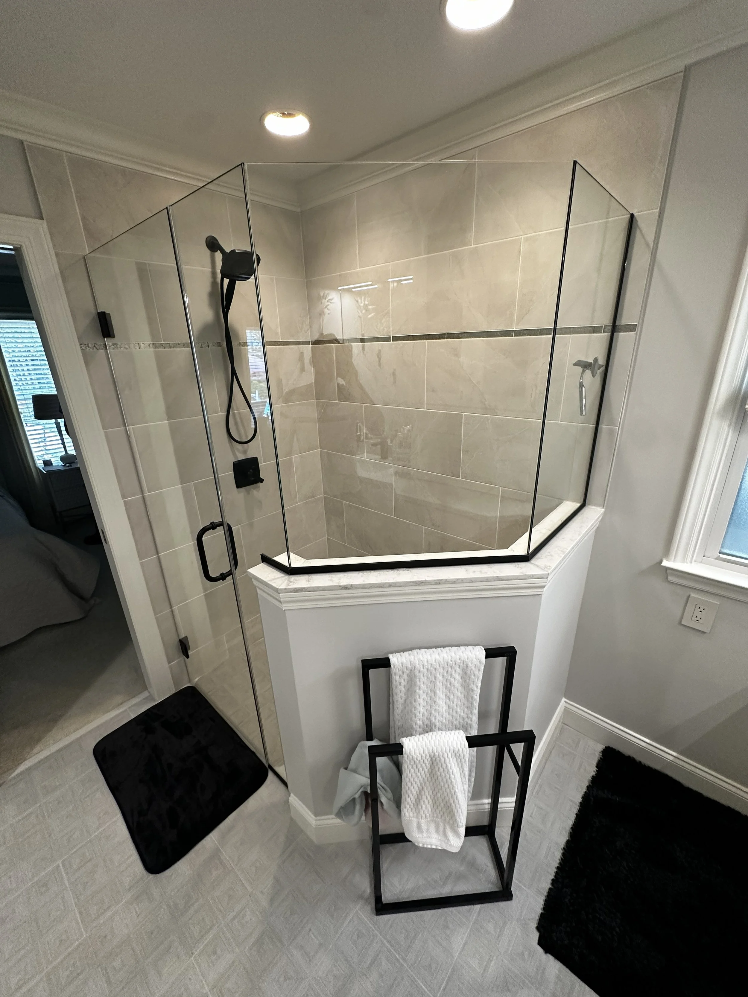 Bathroom shower with glass enclosure, beige tiled walls, and a black towel rack with white towels in front.