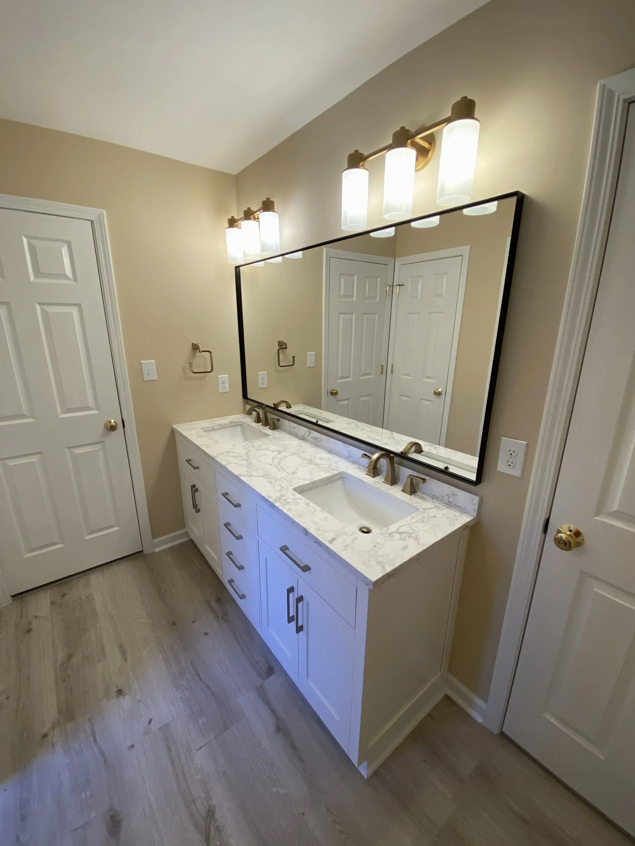 A bathroom vanity with a white marble countertop, two sinks, and a large rectangular mirror. Four light fixtures are mounted above the mirror. The vanity has several drawers and cabinets, and the floor is wooden. There are two closed white doors on adjacent walls.