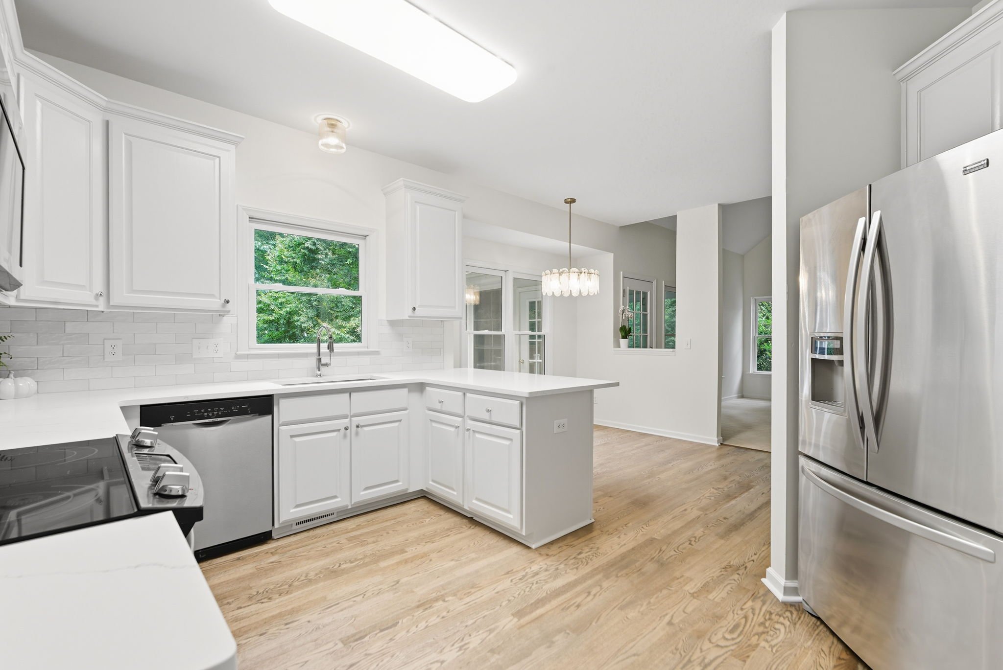 A modern kitchen with white cabinets, a white countertop, and stainless steel appliances. There is a window above the sink and a hanging light fixture over a dining area. The floor is light wood, and the room is filled with natural light.