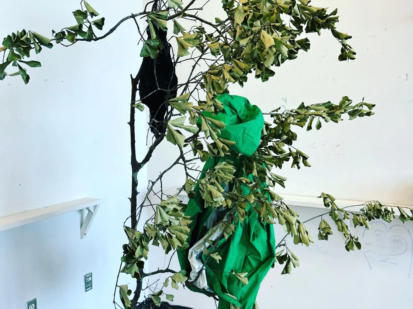 Artificial tree with brown branches and green leaves, some of which are wilting. A green fabric is draped around the lower part of the tree.