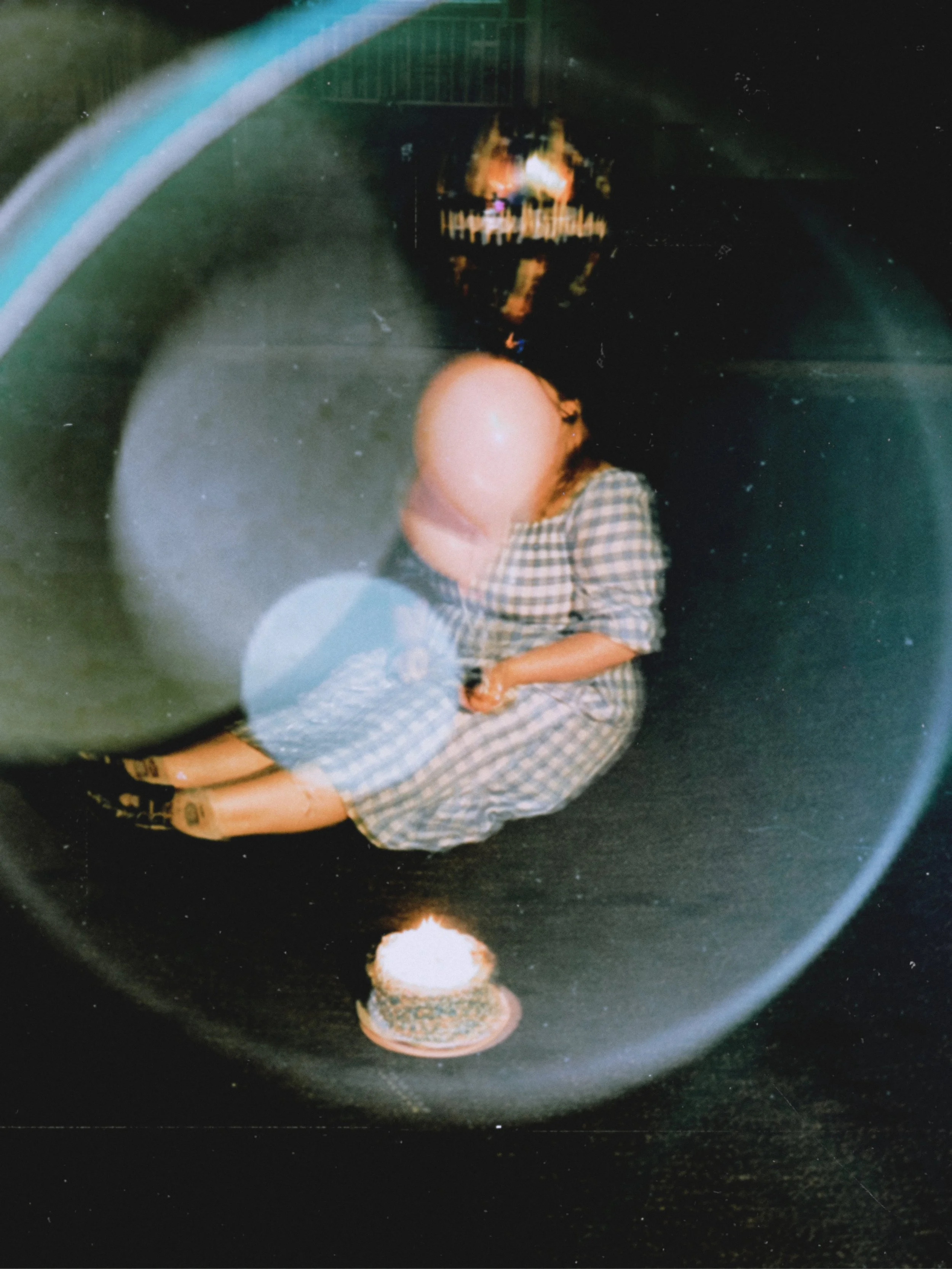 A person with a bald head wearing a checkered dress, sitting in a dark space, with a small birthday cake with a lit candle in front of them, and a creepy skeleton mask reflected in a circular lens or mirror above.
