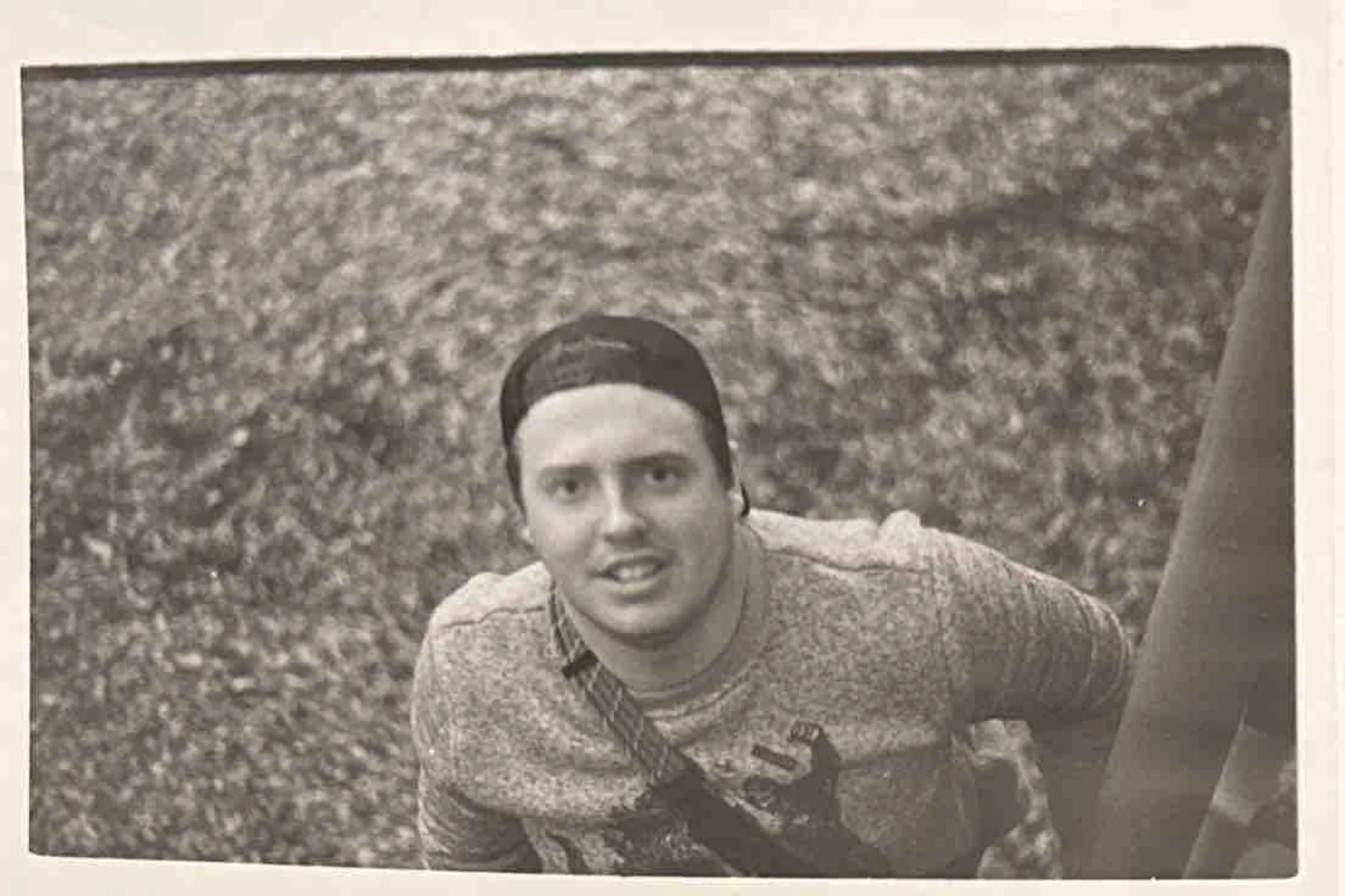 Black and white photo of a young man wearing a cap backwards, looking up at the camera with an intense expression, with a textured surface background.