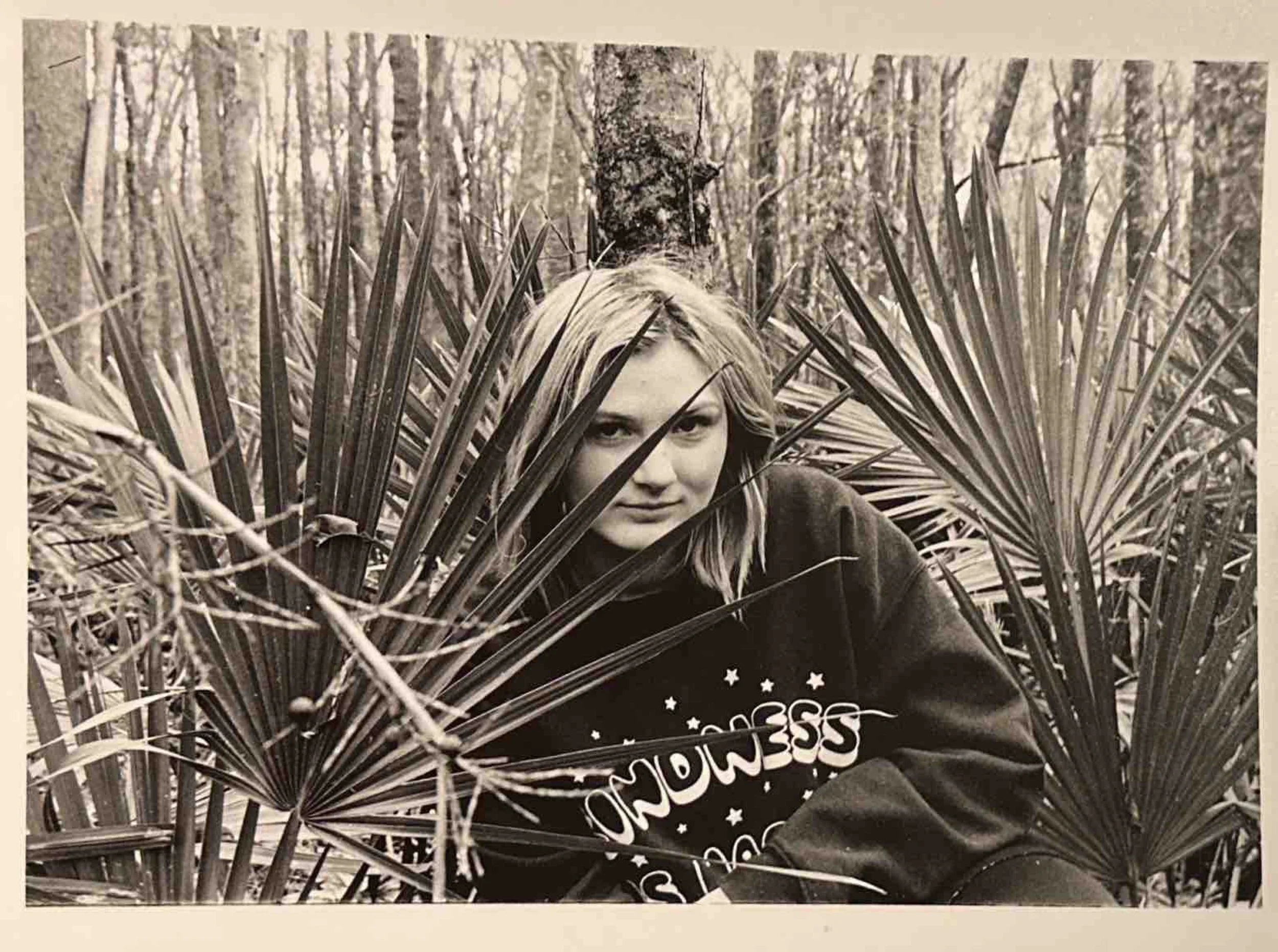 Black and white photo of a young woman with blond hair crouching behind large palm leaves in a forest.