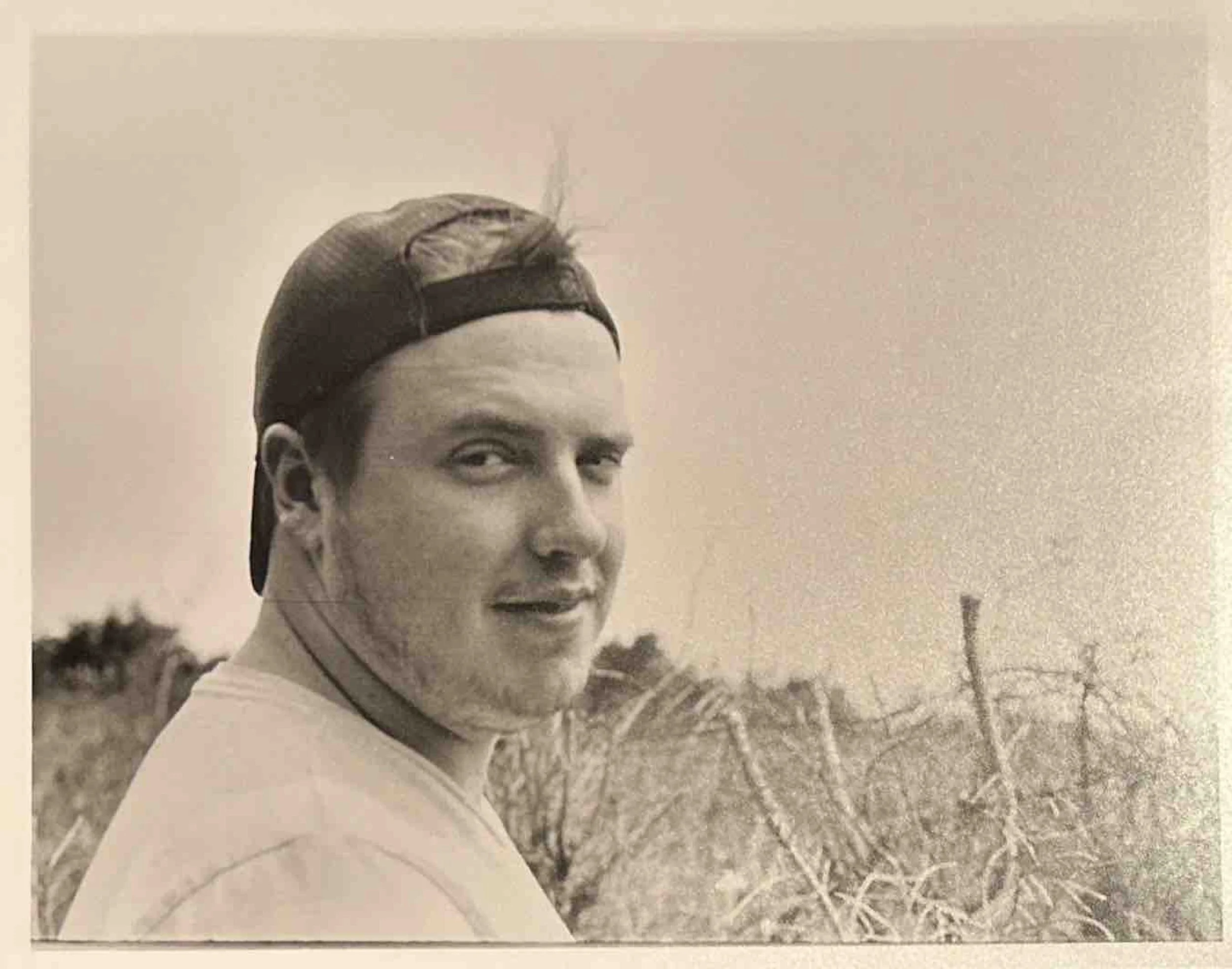 A young man wearing a backward baseball cap, smiling and looking over his shoulder, in an outdoor setting with sparse trees or bushes in the background.