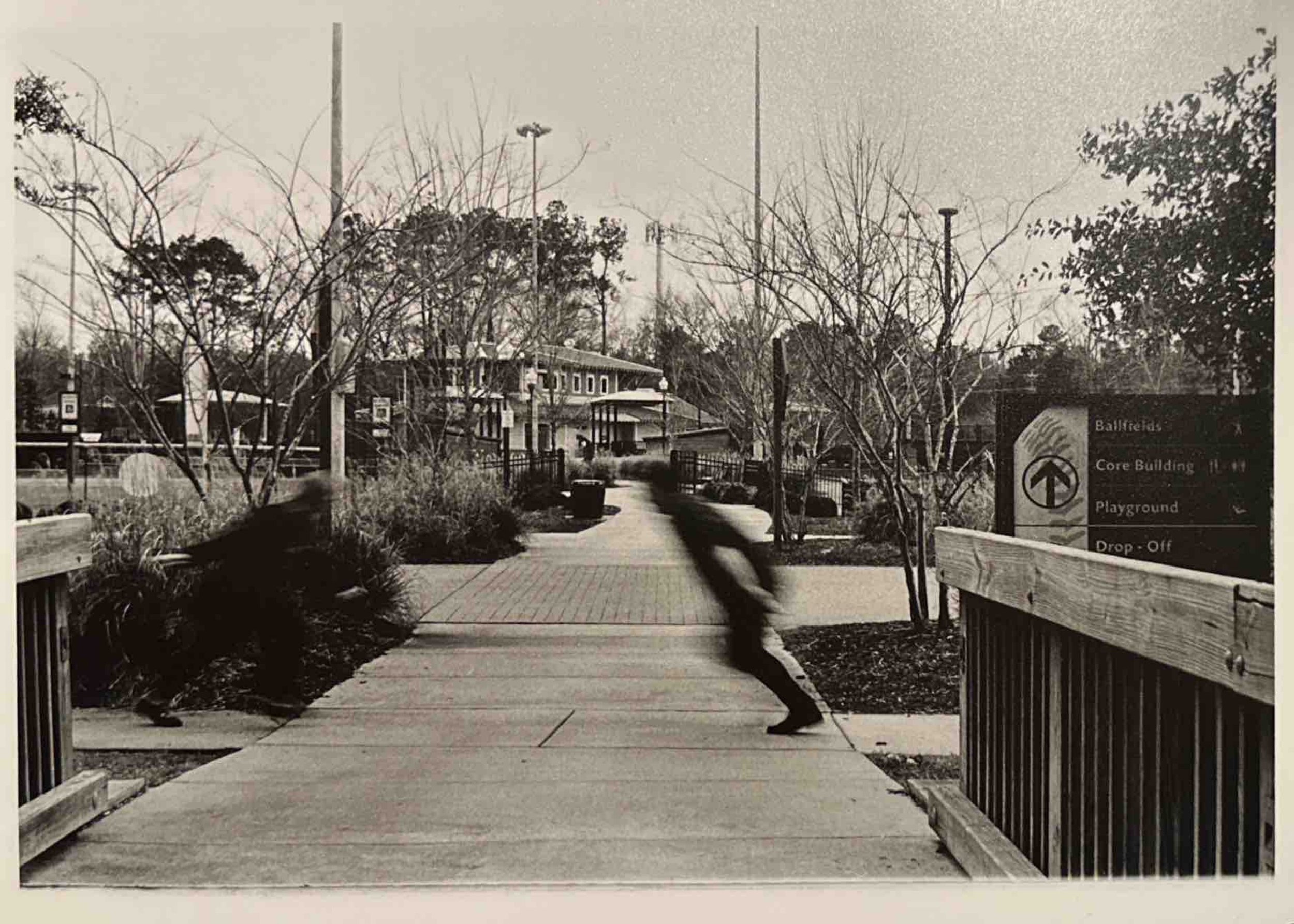 Black and white photo of a park entrance with blurred person walking across the bridge. Sign indicates directions to ballfields, core building, playground, and drop-off.