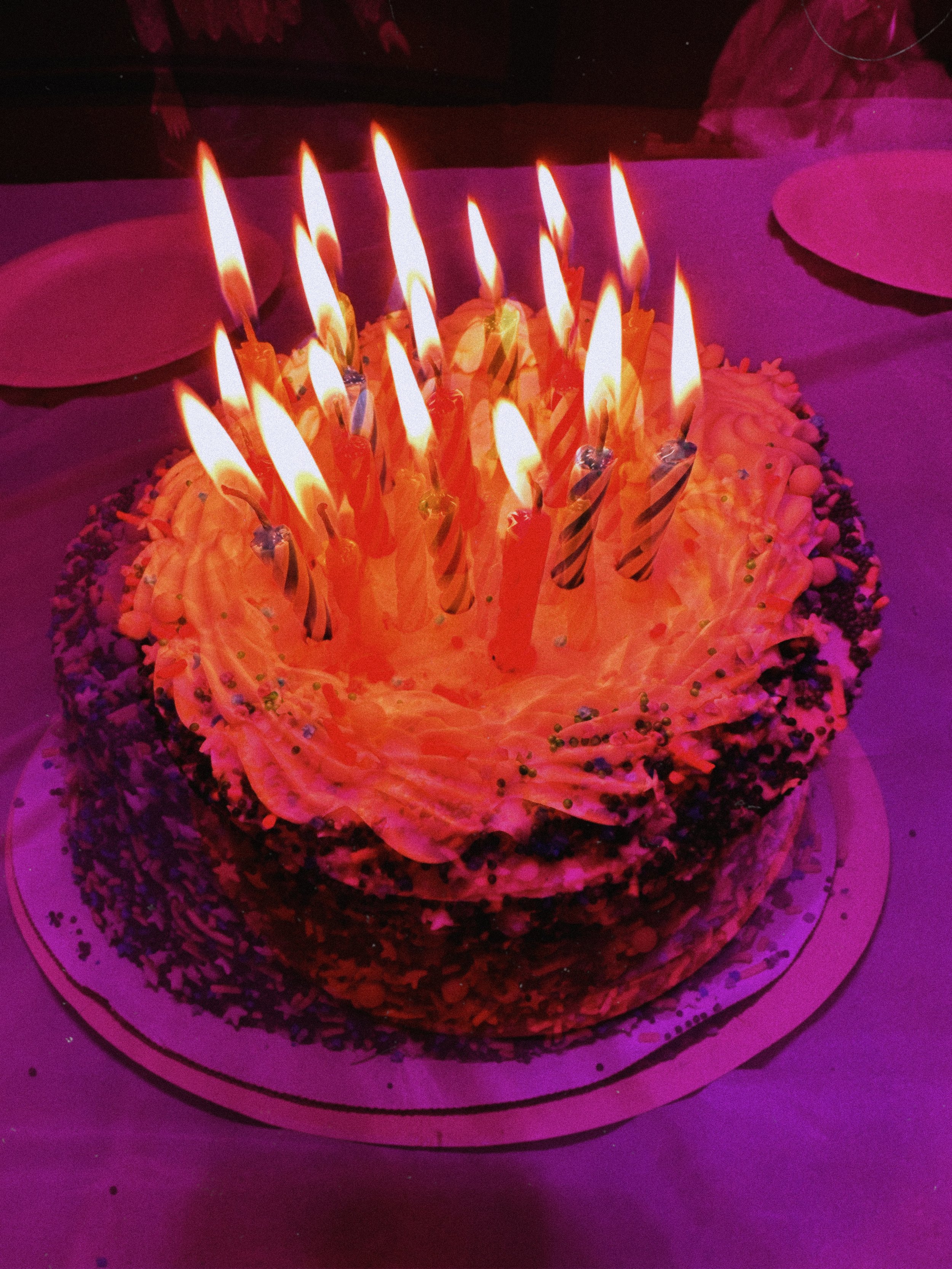 A birthday cake with lit candles on top, decorated with pink frosting, sprinkles, and surrounded by plates.