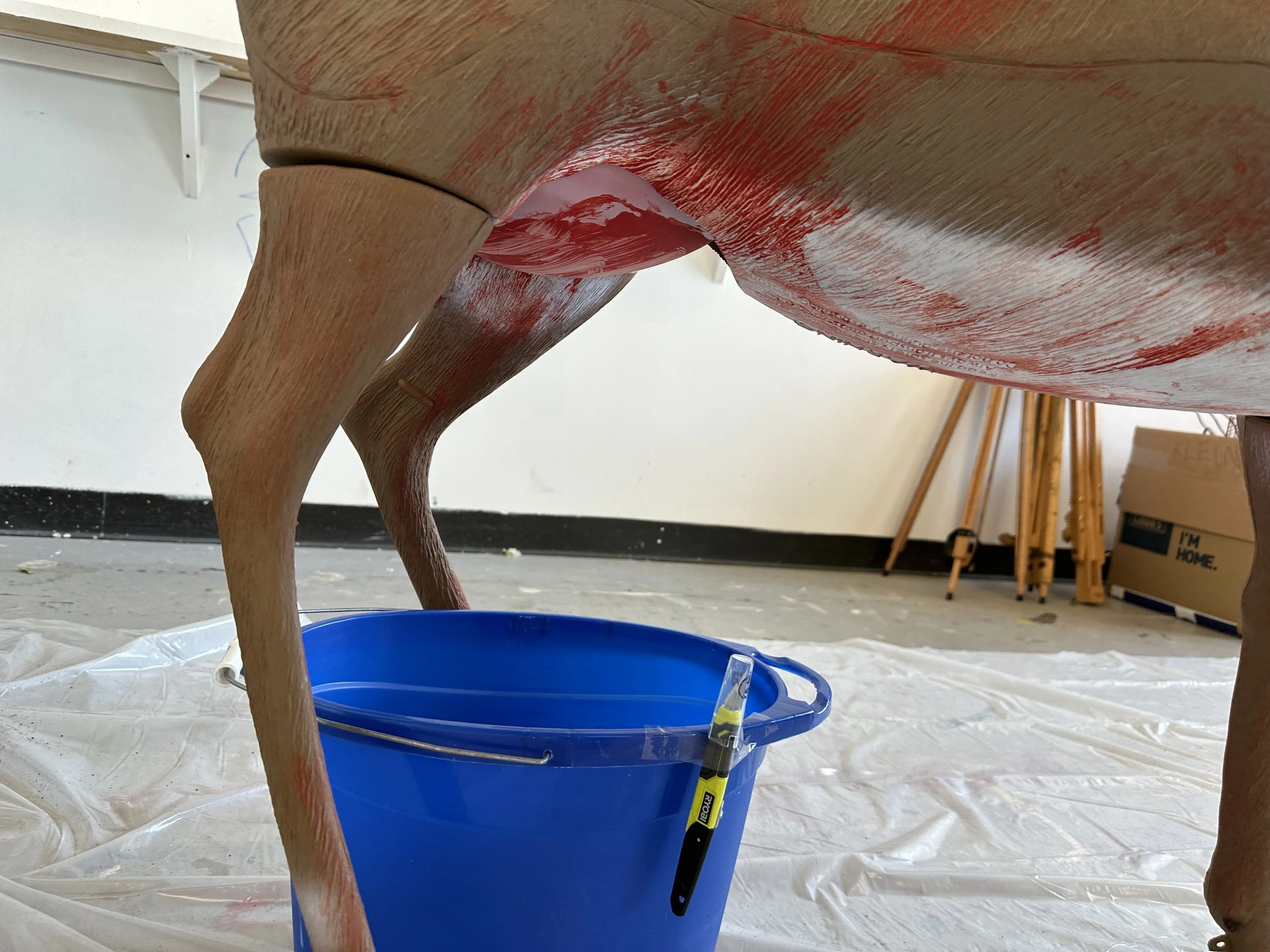 Close-up view of a wooden cow sculpture being painted red, with a blue bucket and paintbrush on the floor underneath, in an indoor workshop.