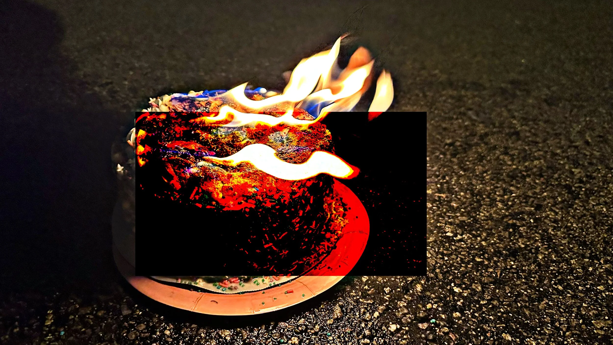 A decorated Christmas cake on a foil-covered plate is on a dark asphalt surface, with flames burning on top of the cake.