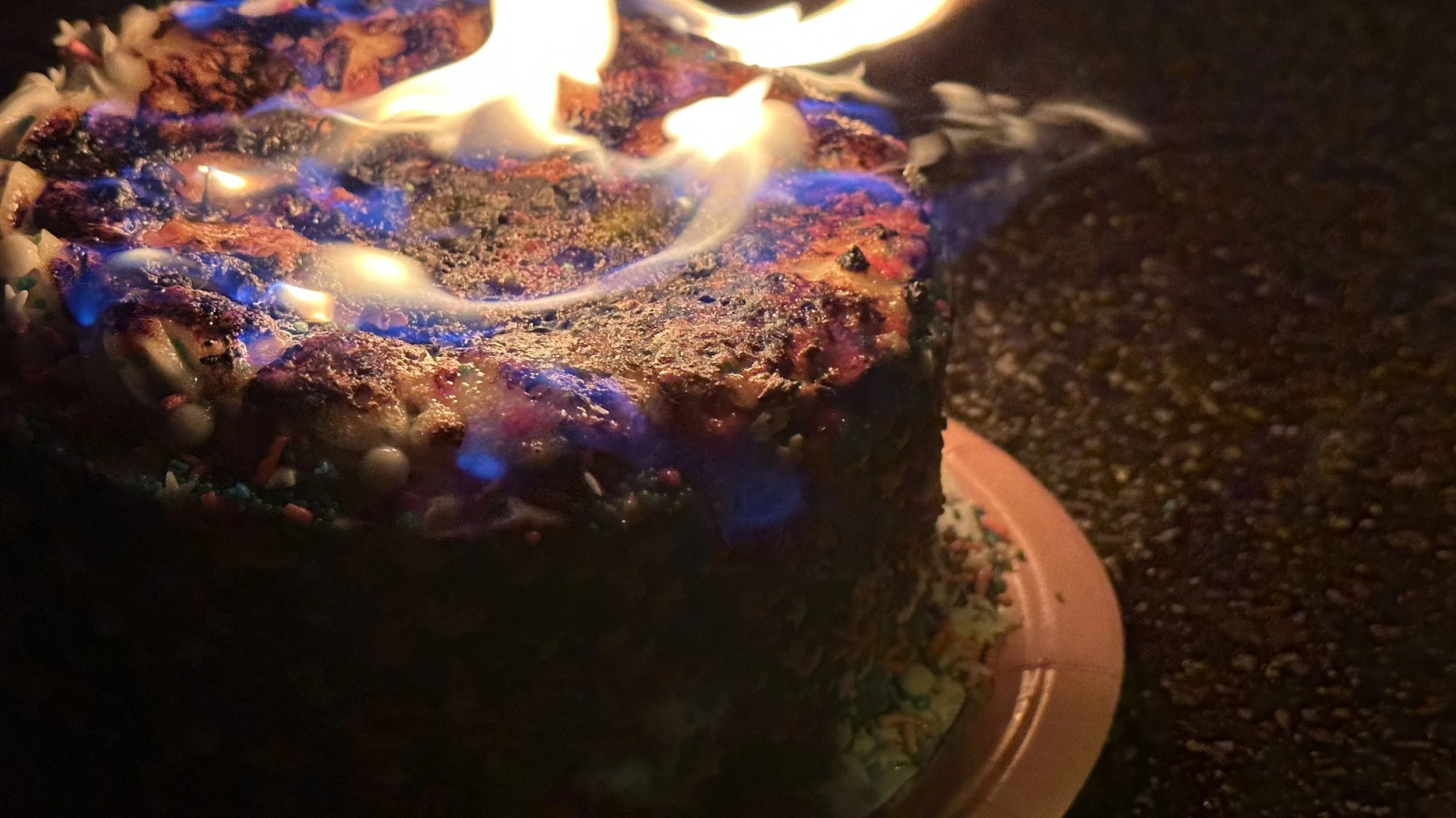 A birthday cake with colorful sprinkles on the sides and frosting on top, set on a plate, with blue and orange flames burning on top in a dark setting.