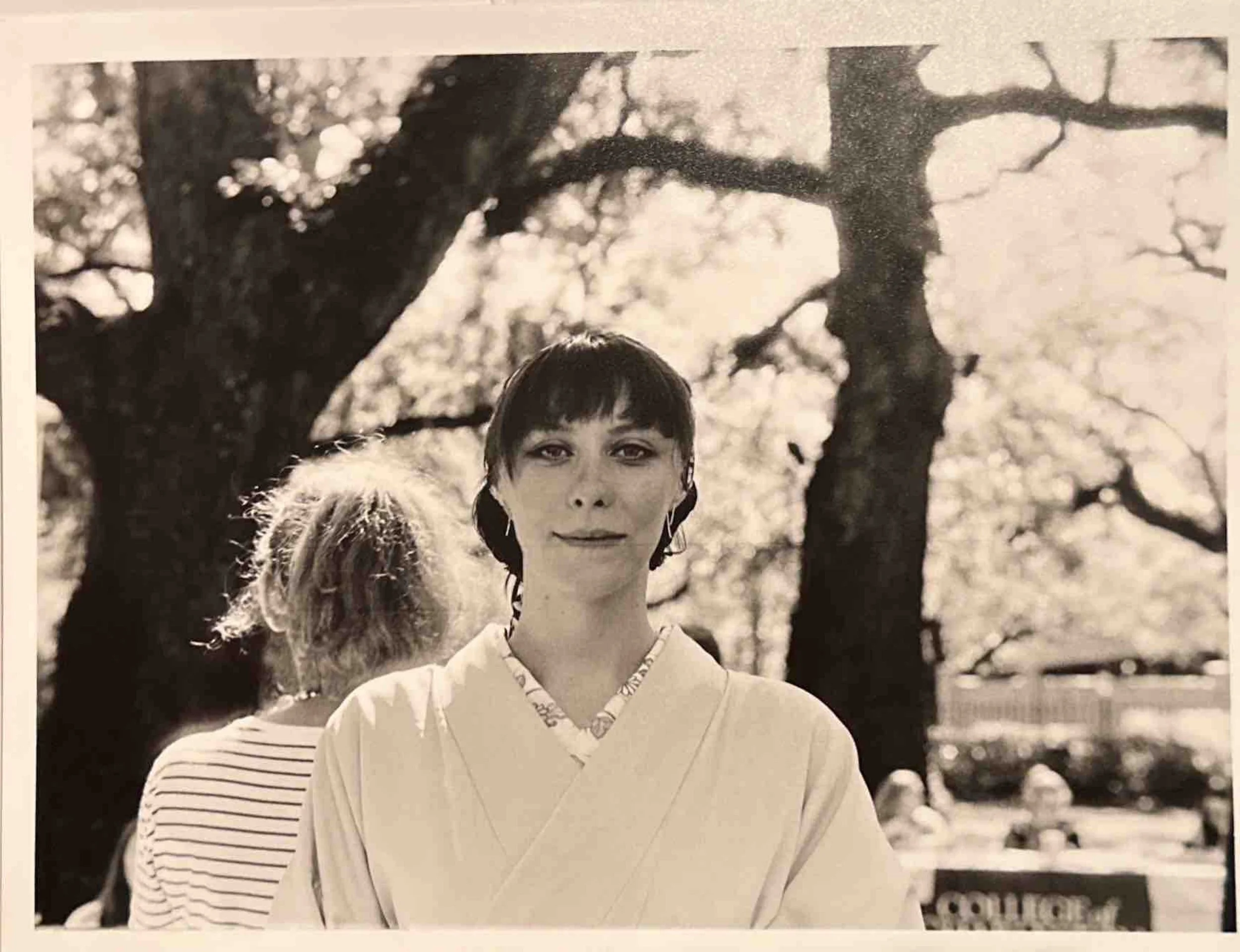 Black and white photo of a woman in traditional attire standing outdoors with large trees and a fence in the background, and a person with curly hair in a striped shirt behind her.