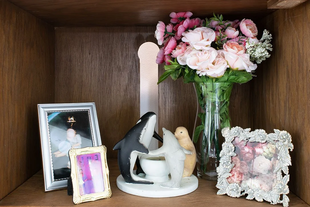 A wooden shelf with a framed photograph, a smaller ornate photo frame, ceramic dolphin and seal figurines around a white bowl, and a tall glass vase with pink and white flowers.